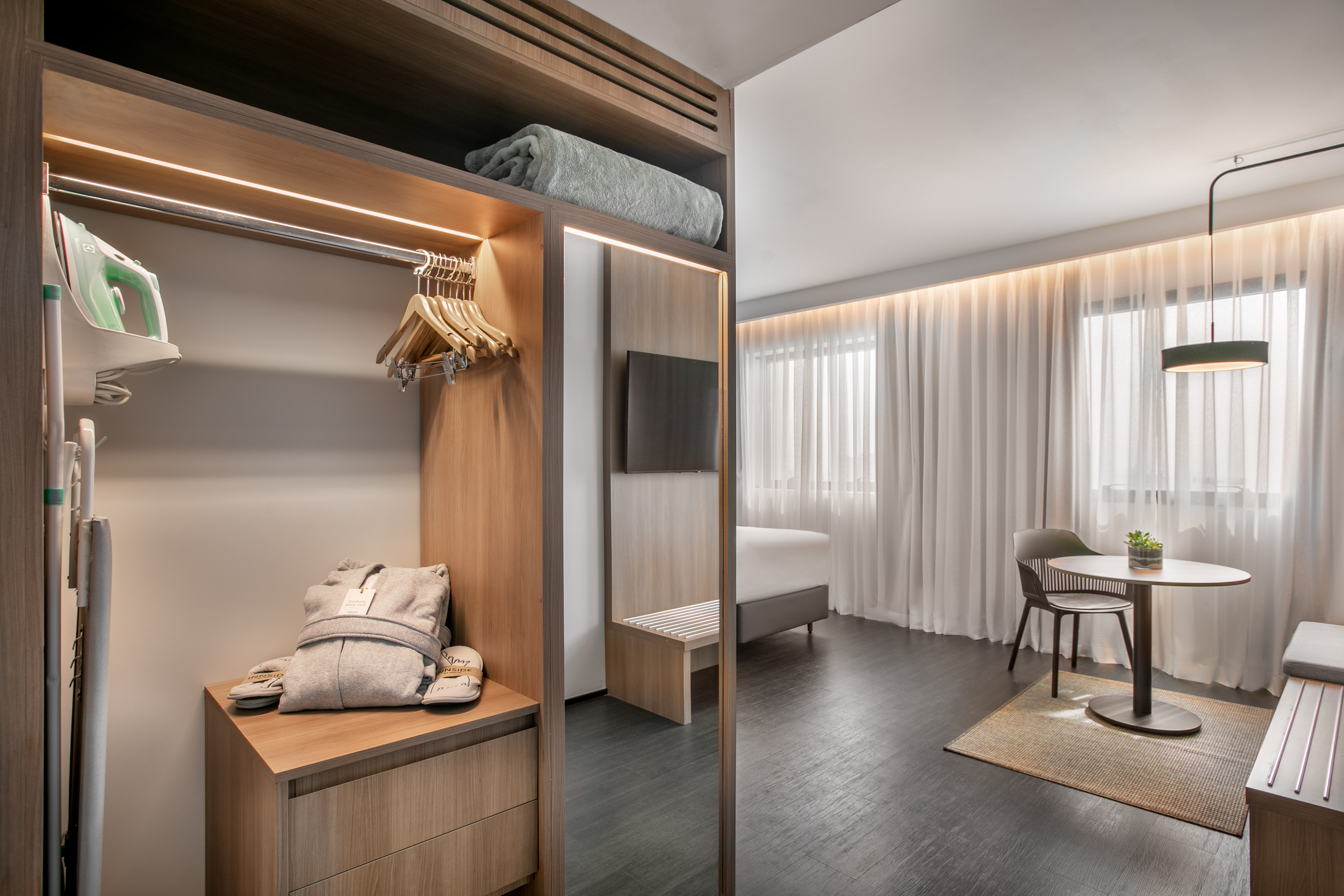 Hotel room with open wardrobe, iron, bathrobe, slippers, bed, and dining area.