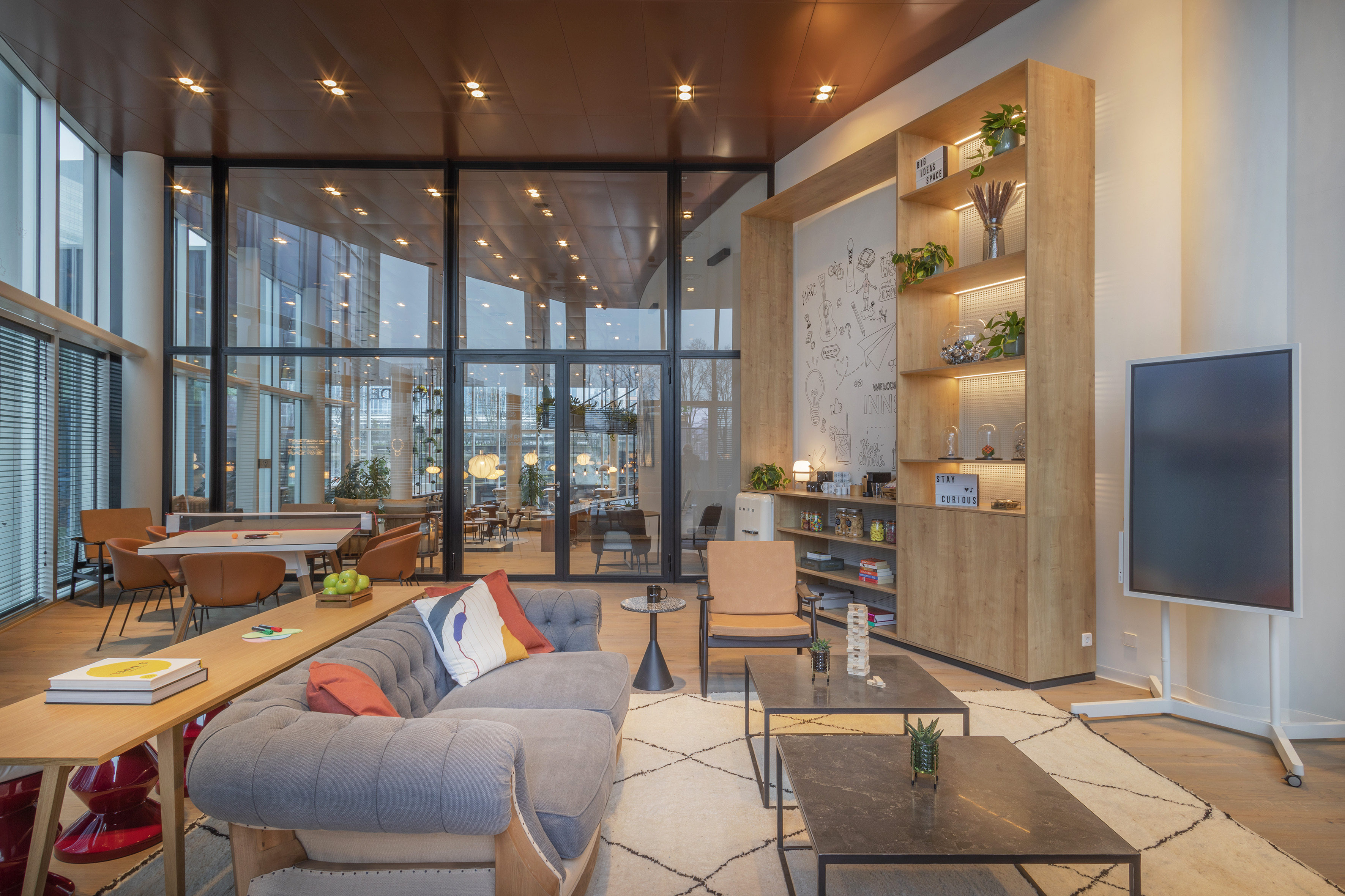 Modern office lounge interior with grey sofa, wood shelving, and large windows.