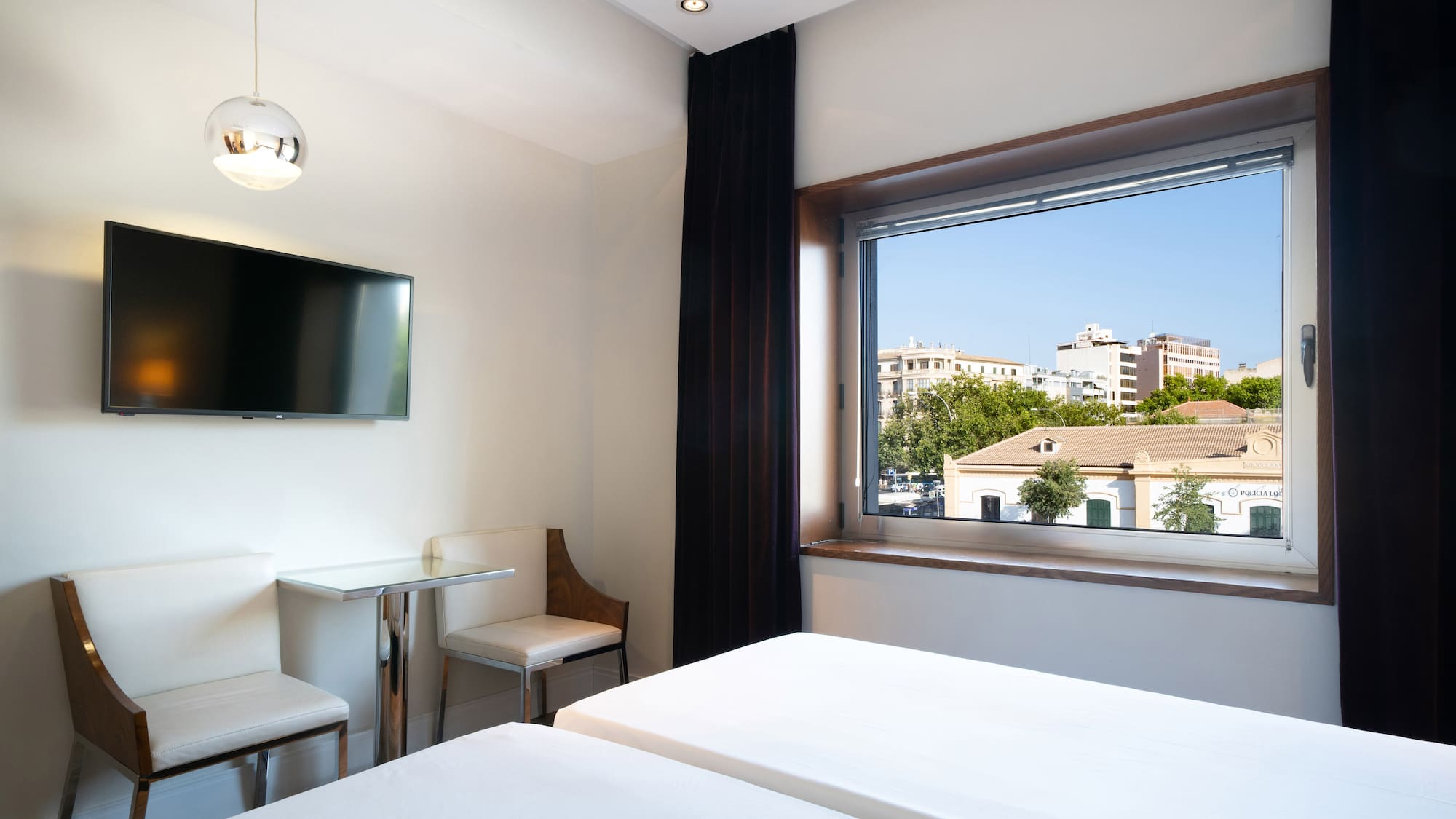 Modern hotel room with twin beds, wall TV, city view window, and seating area.