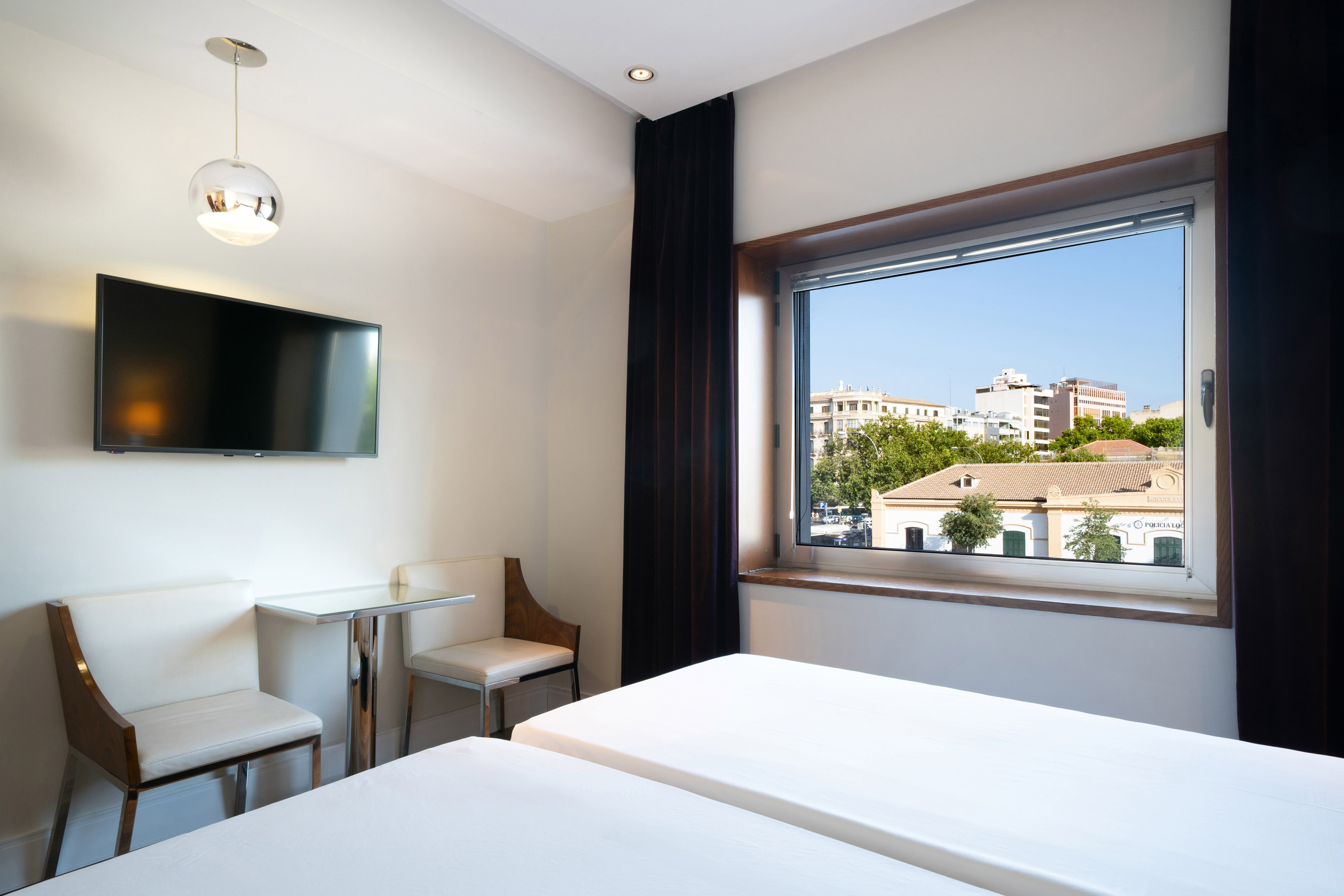 Modern hotel room with twin beds, wall TV, city view window, and seating area.