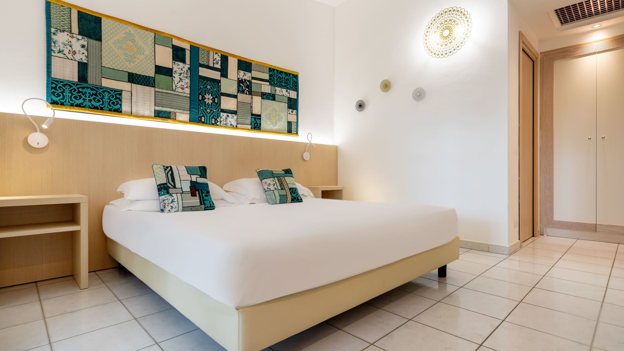 Modern hotel bedroom with double bed, vibrant textile wall art, and tiled floor.