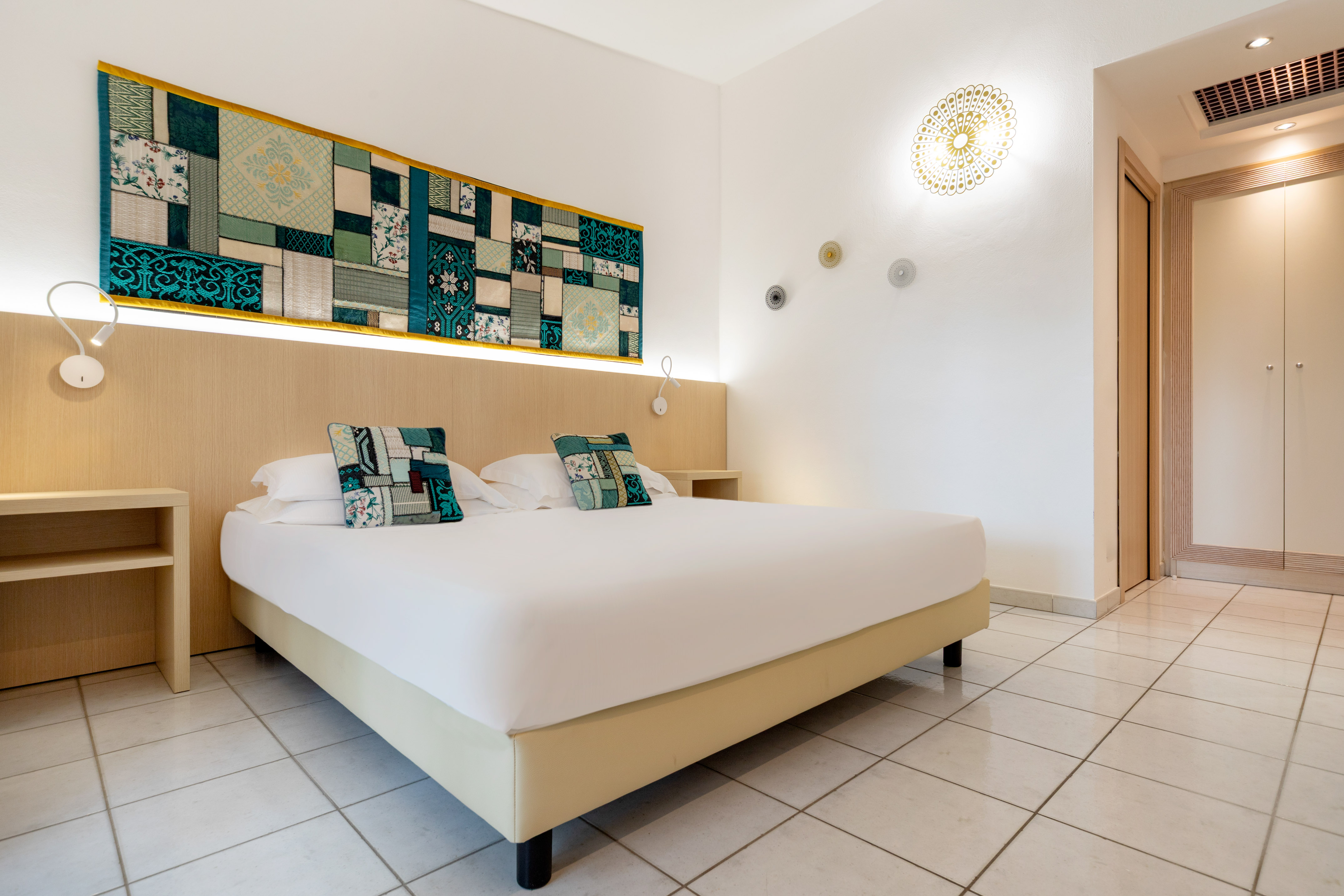Modern hotel bedroom with double bed, vibrant textile wall art, and tiled floor.