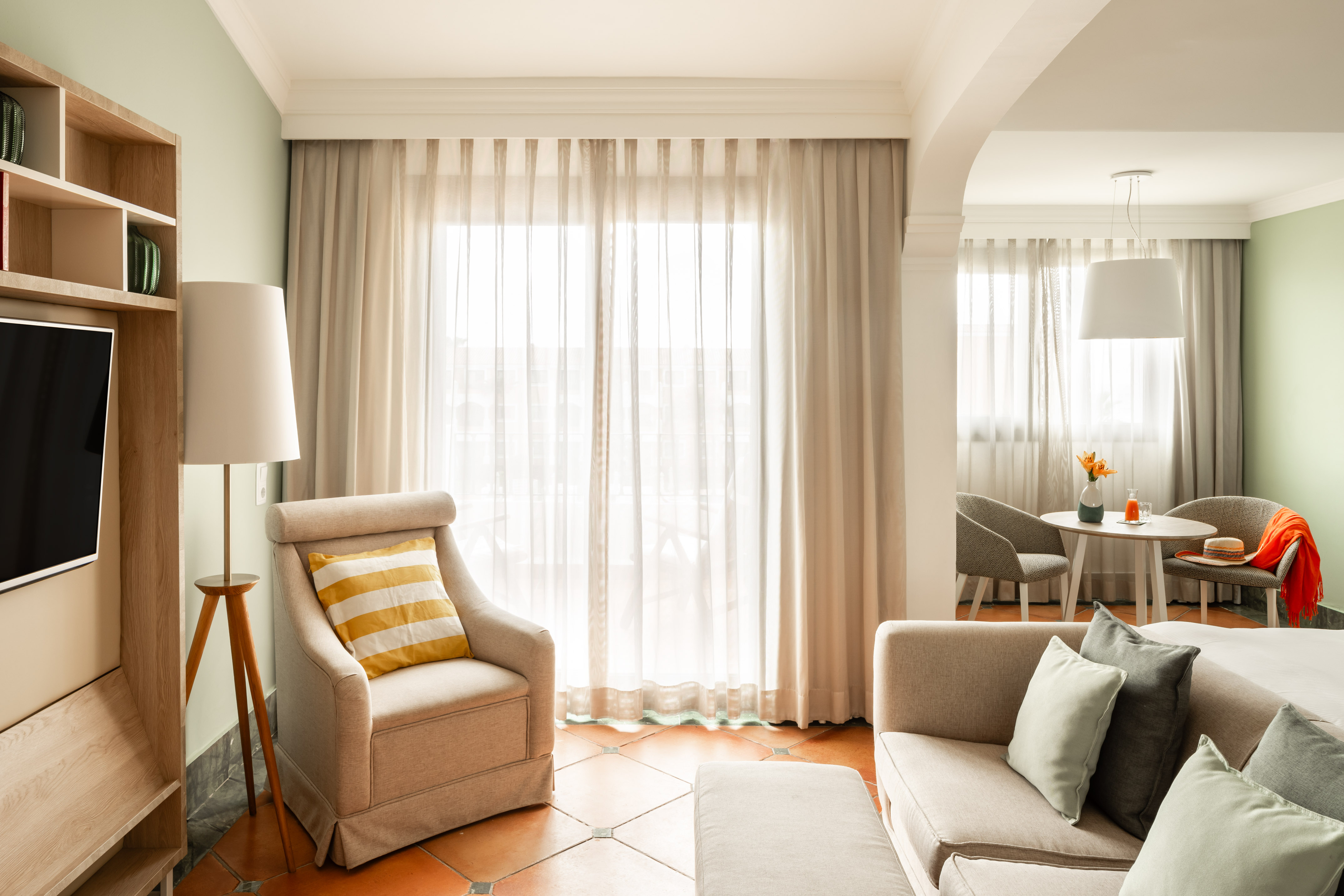Sunlit hotel suite living room with modern sofa, armchair, TV, and dining area.