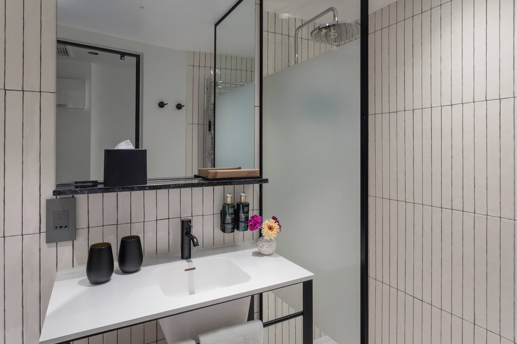 Modern bathroom with white vertical tiles, black vanity, rainfall shower, and mirror.
