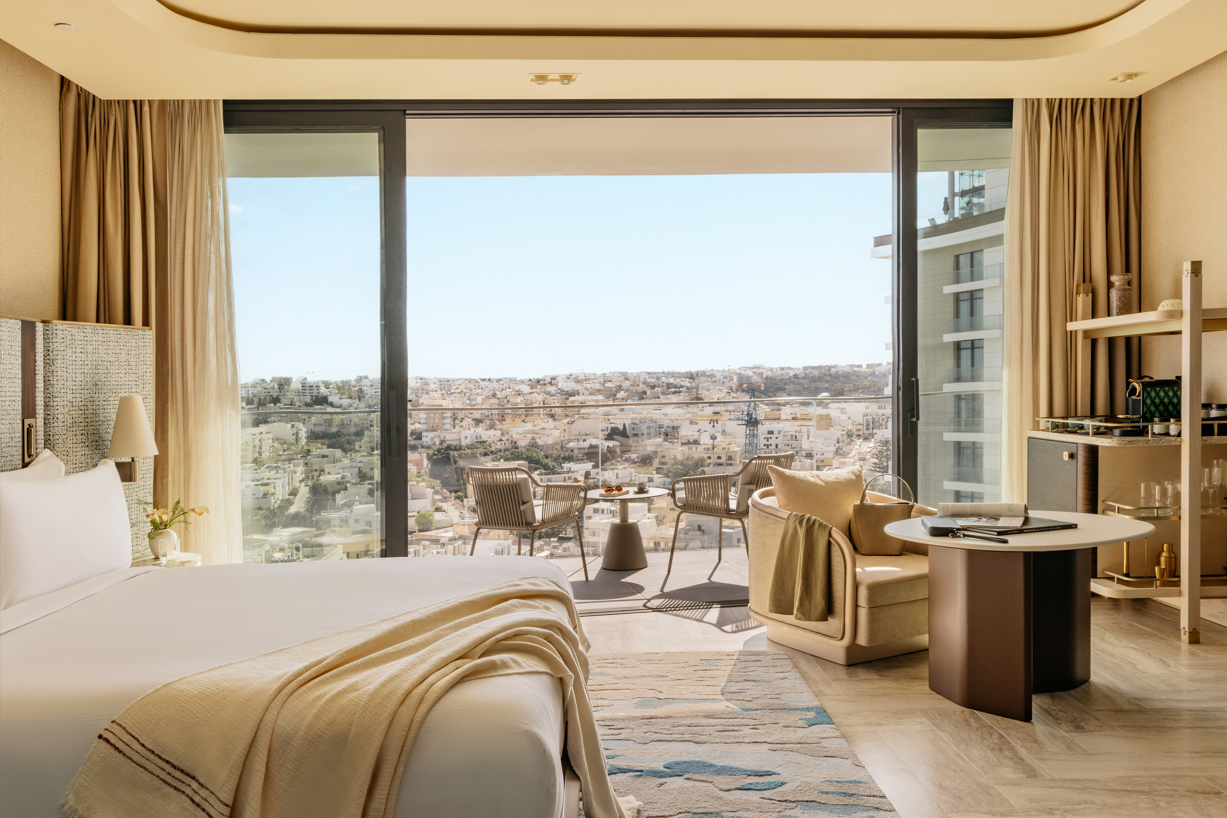 Luxury hotel room with balcony, city view, bed, and outdoor seating.
