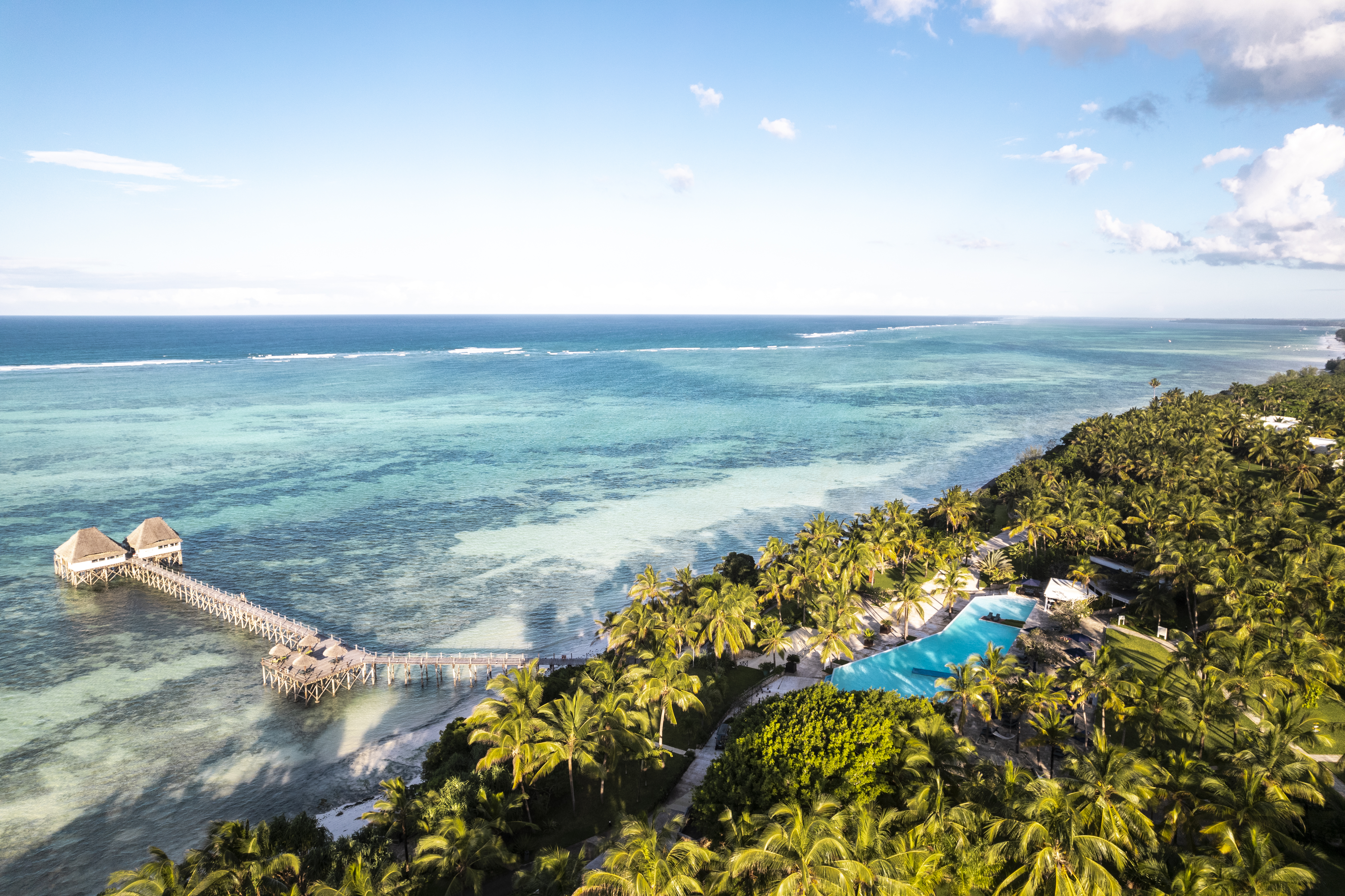 Aerial view of tropical ocean resort with pier, palm trees, and swimming pool.