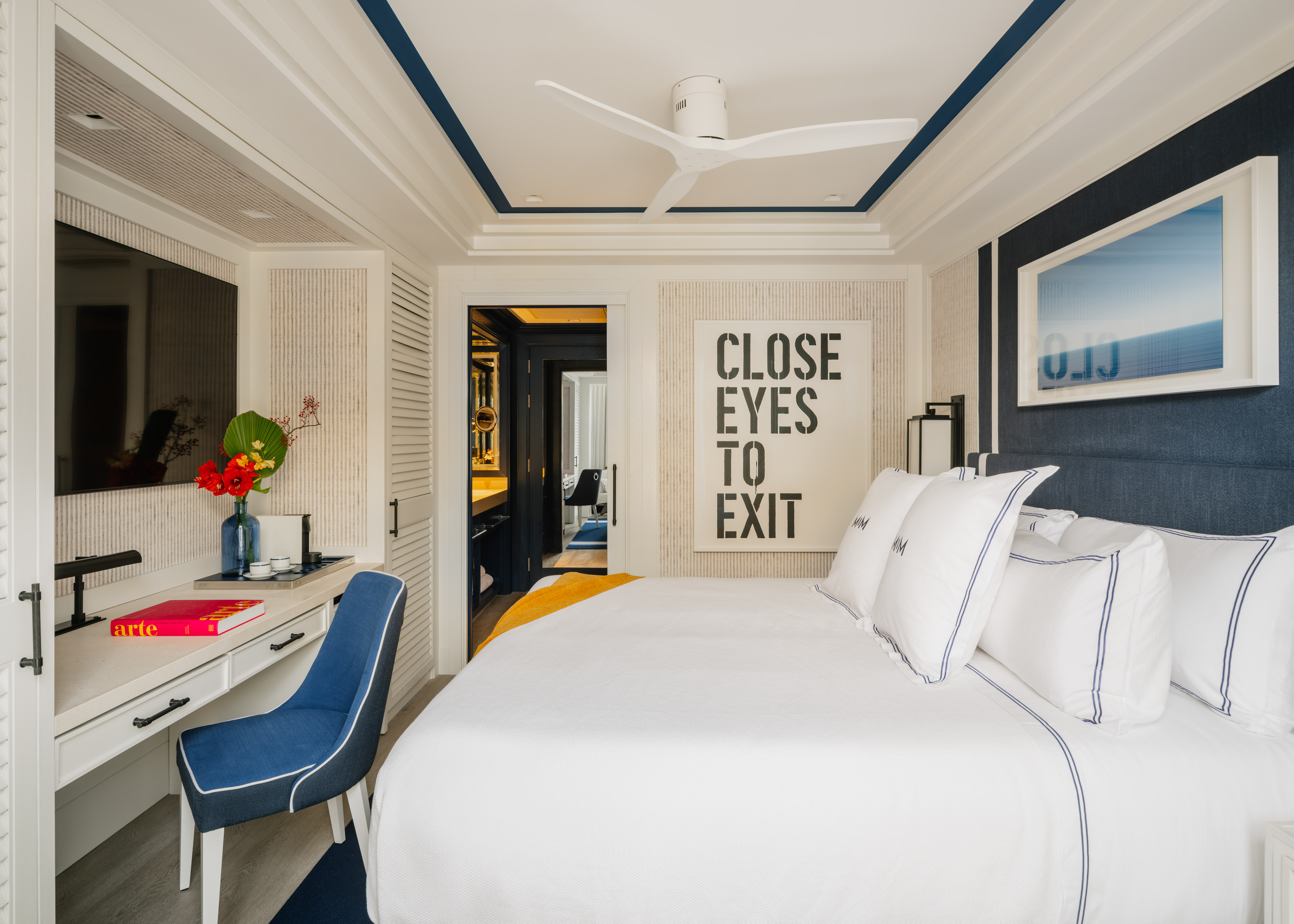 Luxury hotel bedroom with white bed, blue accents, desk, and unique art piece.
