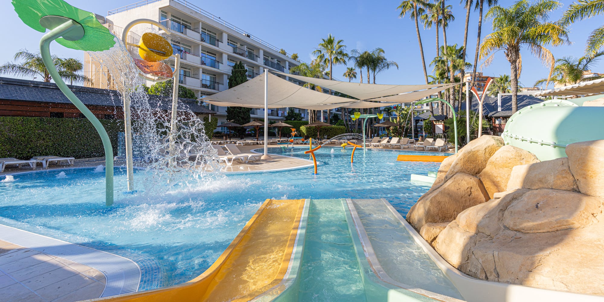 Sunny hotel water park with slides, splashing play area, pool, and palm trees.