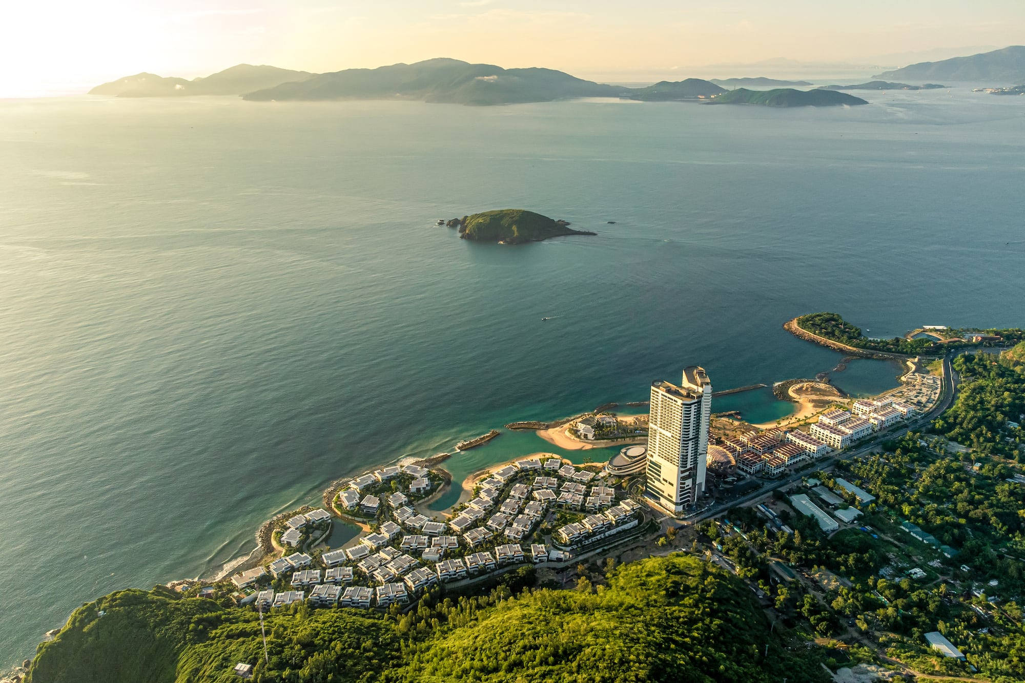 Aerial of coastal resort, high-rise hotel, villas, ocean, and green mountains.