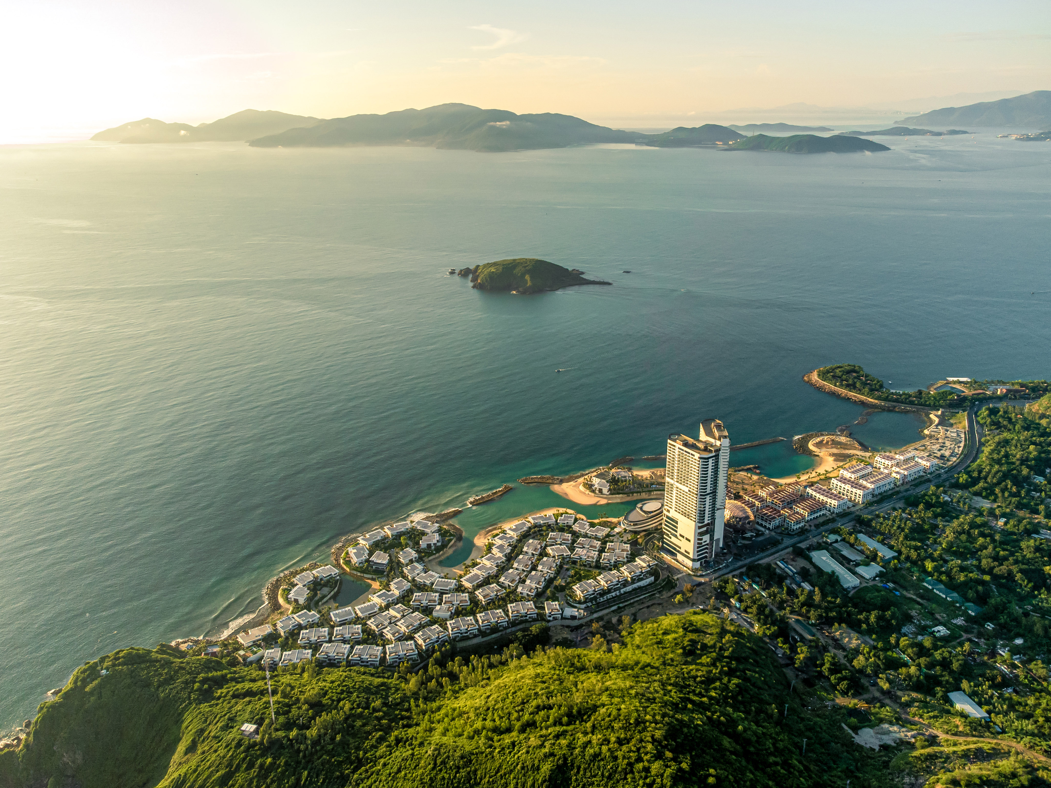 Aerial of coastal resort, high-rise hotel, villas, ocean, and green mountains.
