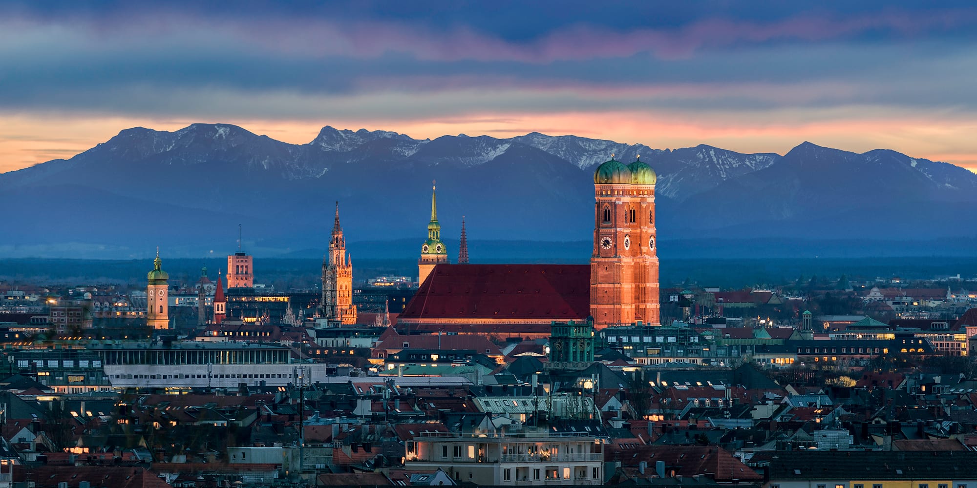 Múnich al atardecer: Frauenkirche, luces urbanas y Alpes nevados.
