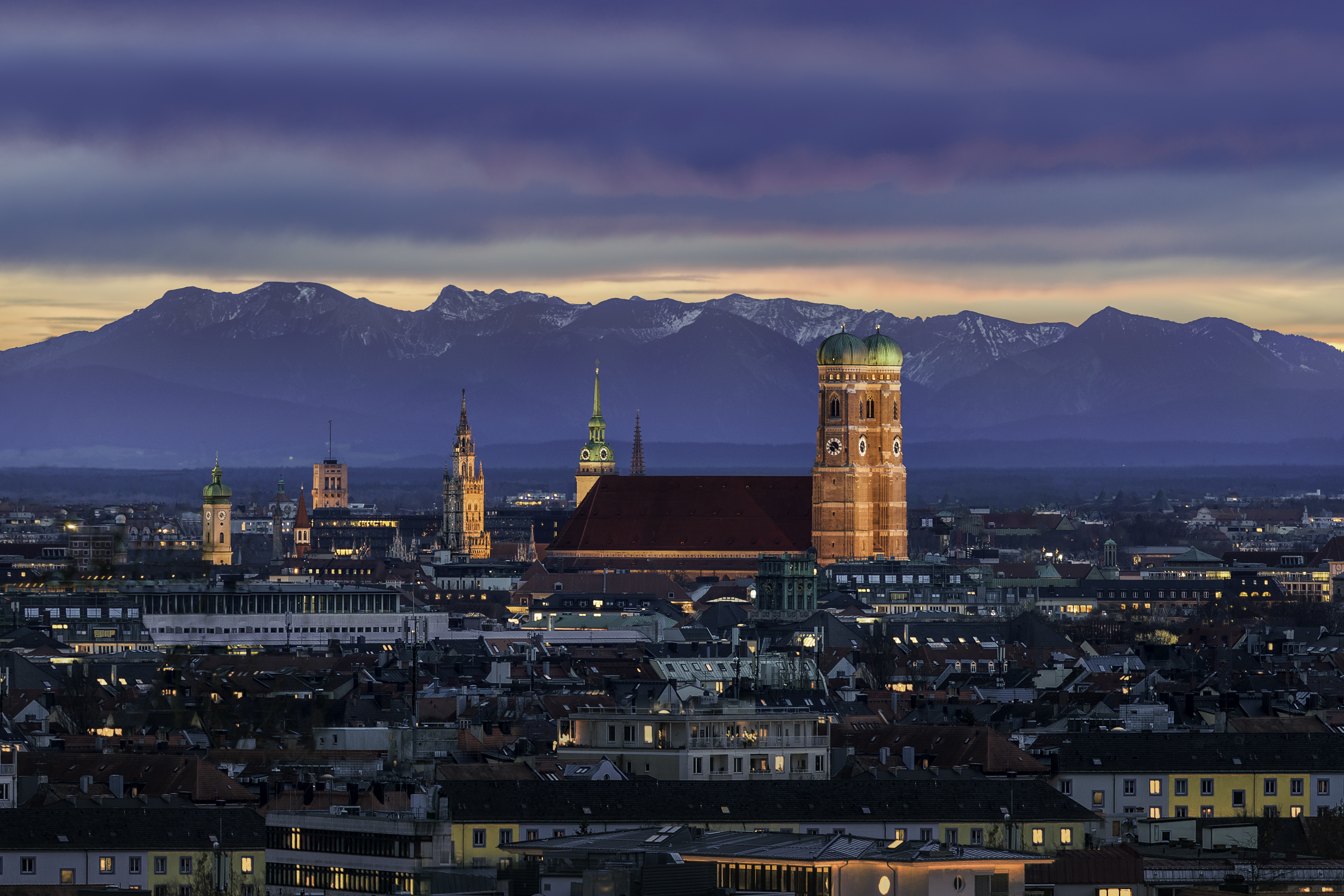 Múnich al atardecer: Frauenkirche, luces urbanas y Alpes nevados.