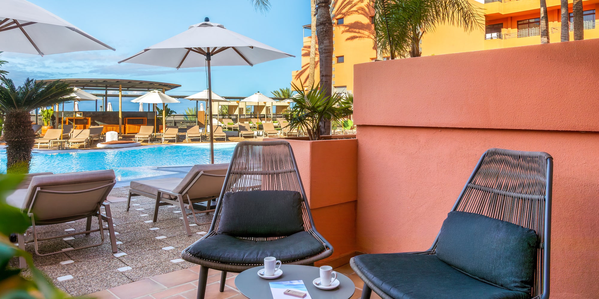 Sunny hotel balcony with chairs, coffee, overlooking resort pool and palm trees.