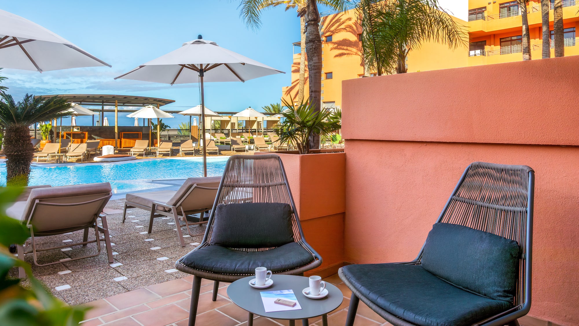 Sunny hotel balcony with chairs, coffee, overlooking resort pool and palm trees.