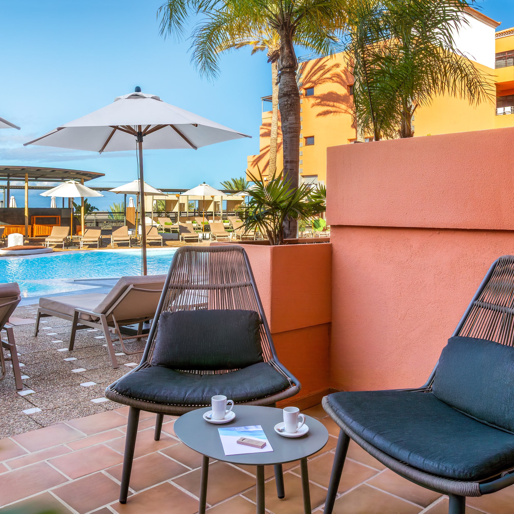 Sunny hotel balcony with chairs, coffee, overlooking resort pool and palm trees.