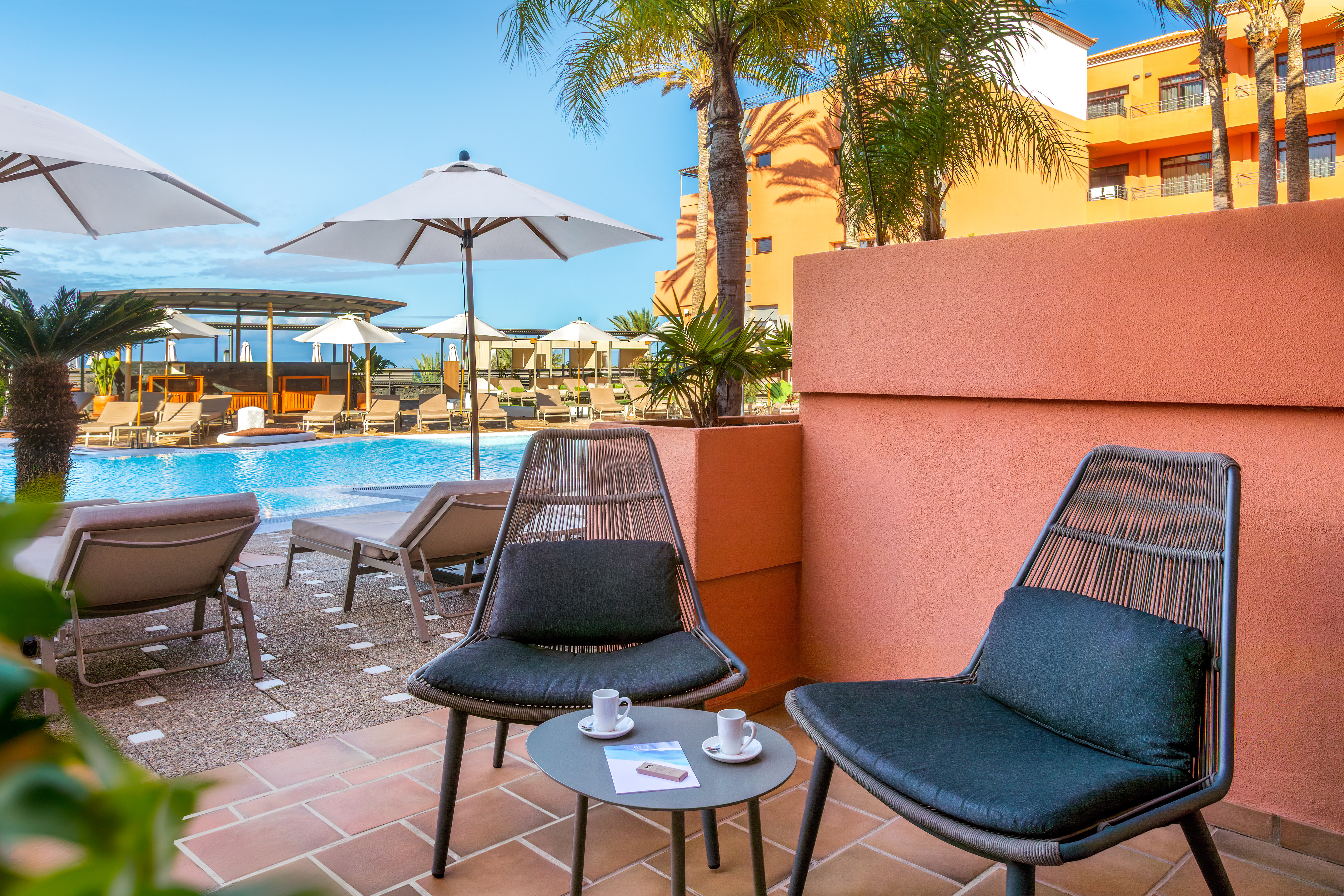 Sunny hotel balcony with chairs, coffee, overlooking resort pool and palm trees.