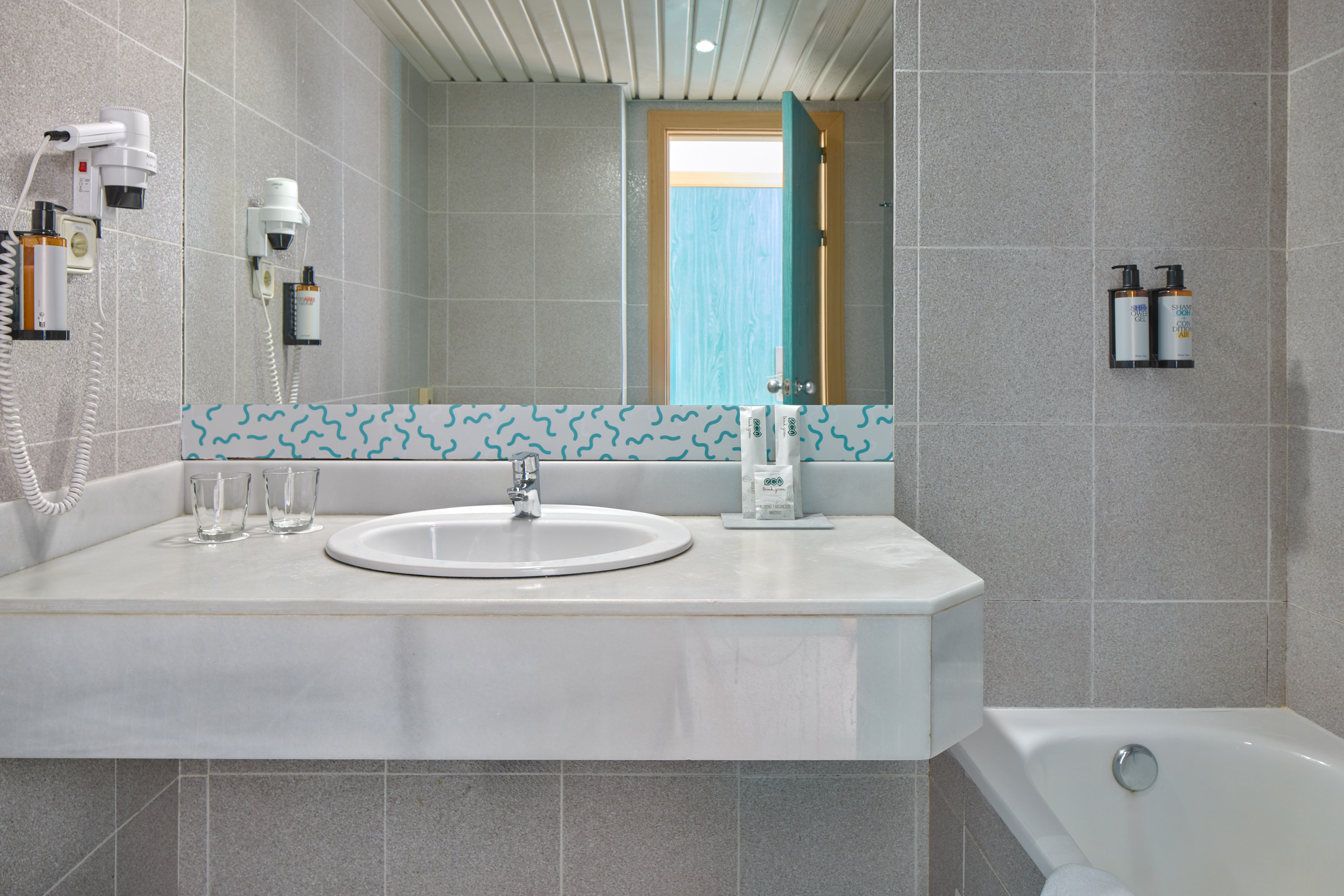 Gray tiled hotel bathroom with white vanity, sink, mirror, hairdryer, and bathtub.
