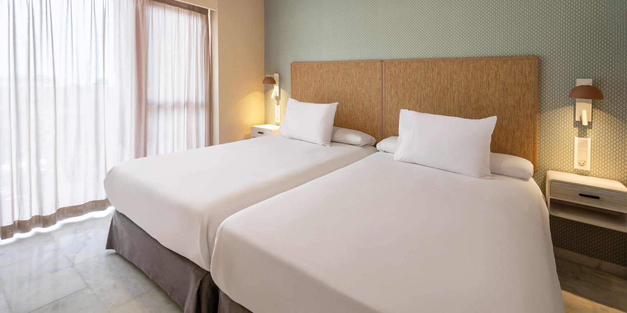 Twin beds in hotel room with woven headboard, patterned wall, and sheer curtains.