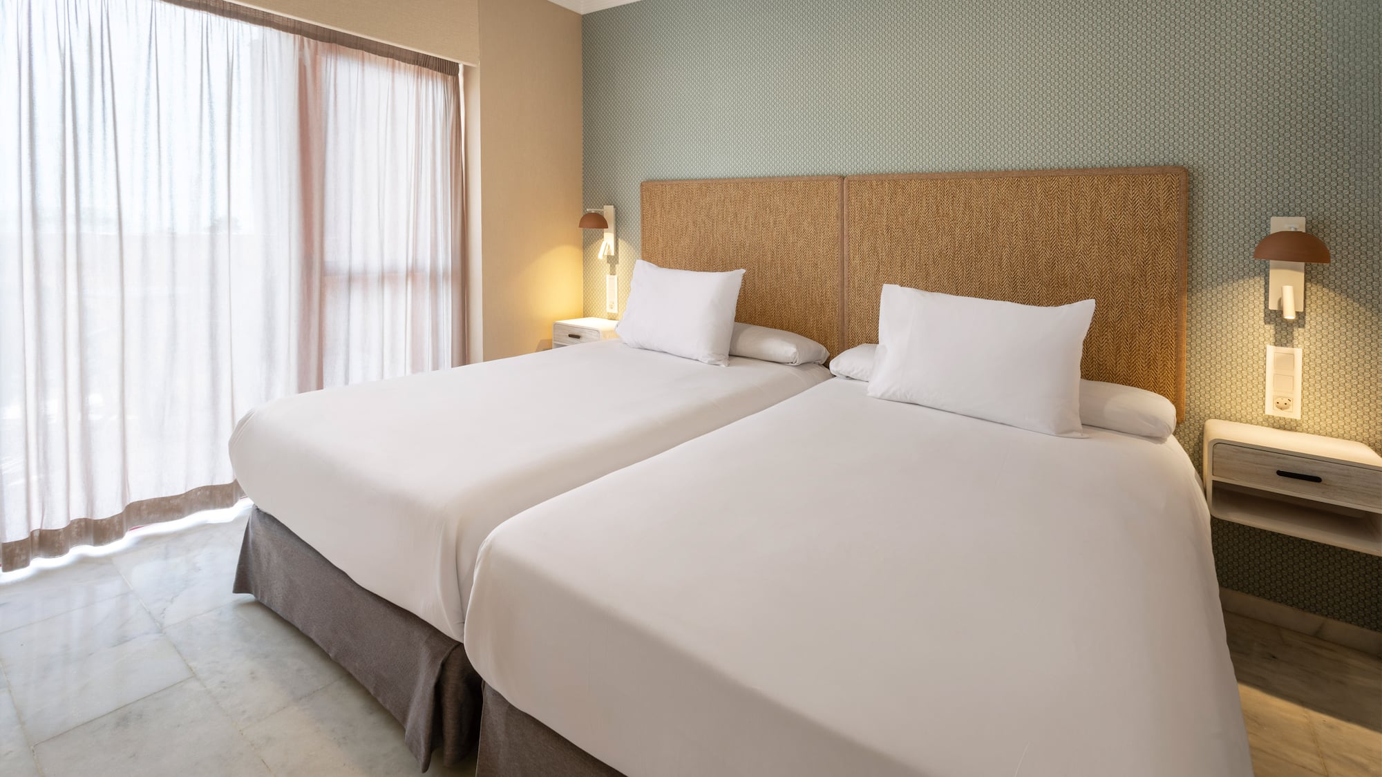 Twin beds in hotel room with woven headboard, patterned wall, and sheer curtains.