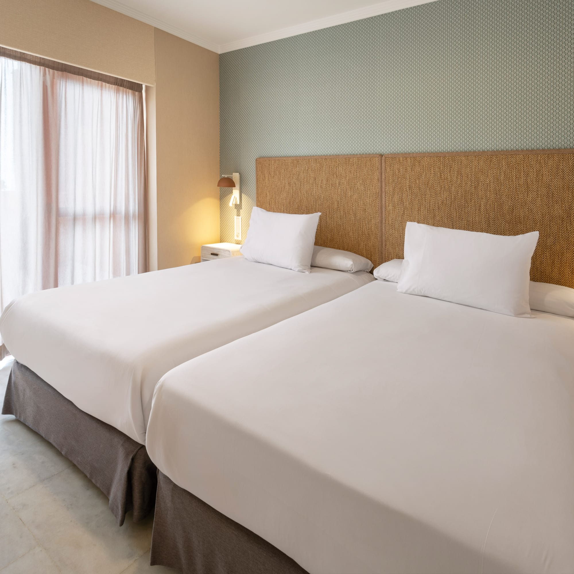 Twin beds in hotel room with woven headboard, patterned wall, and sheer curtains.