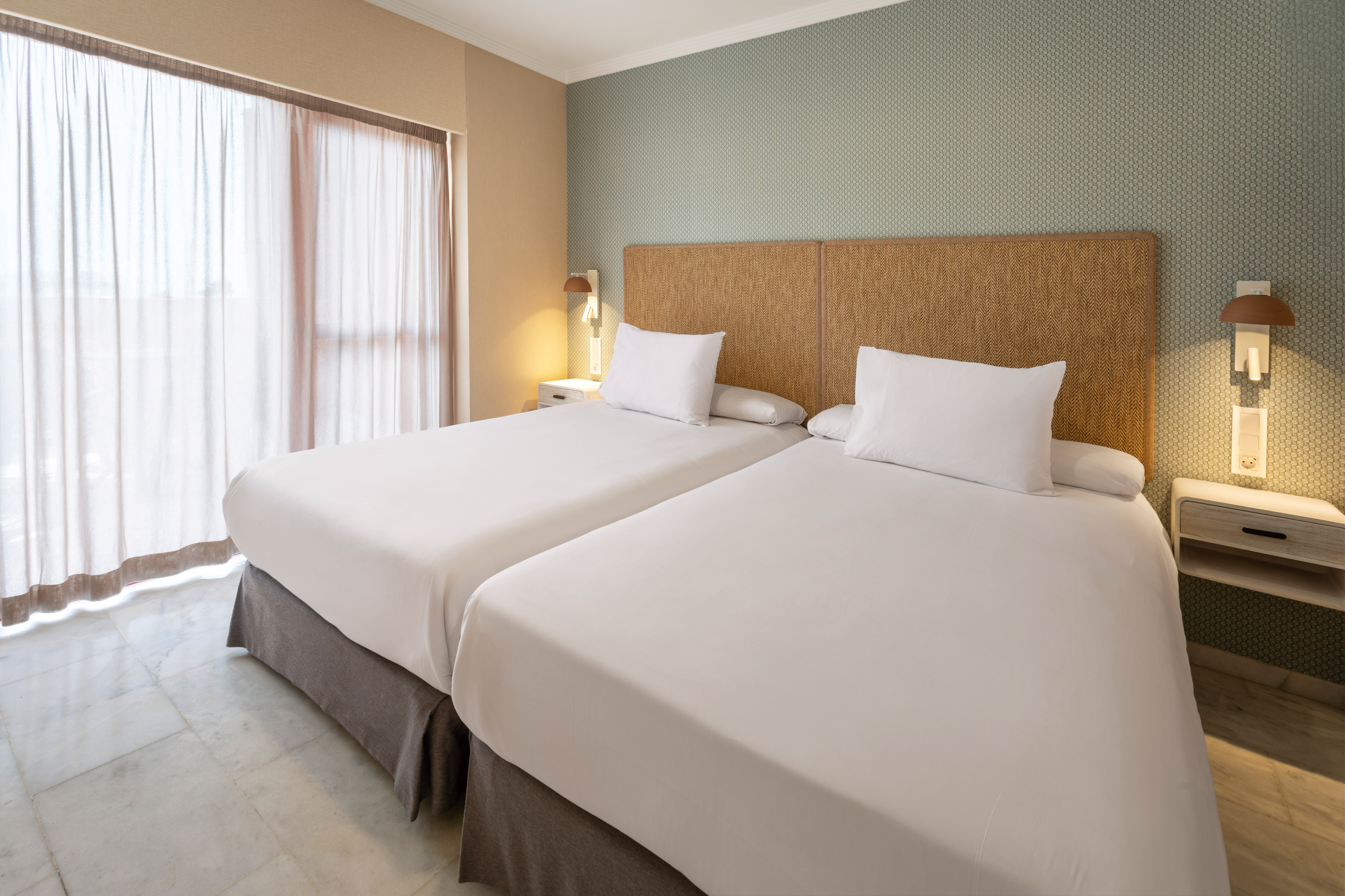 Twin beds in hotel room with woven headboard, patterned wall, and sheer curtains.
