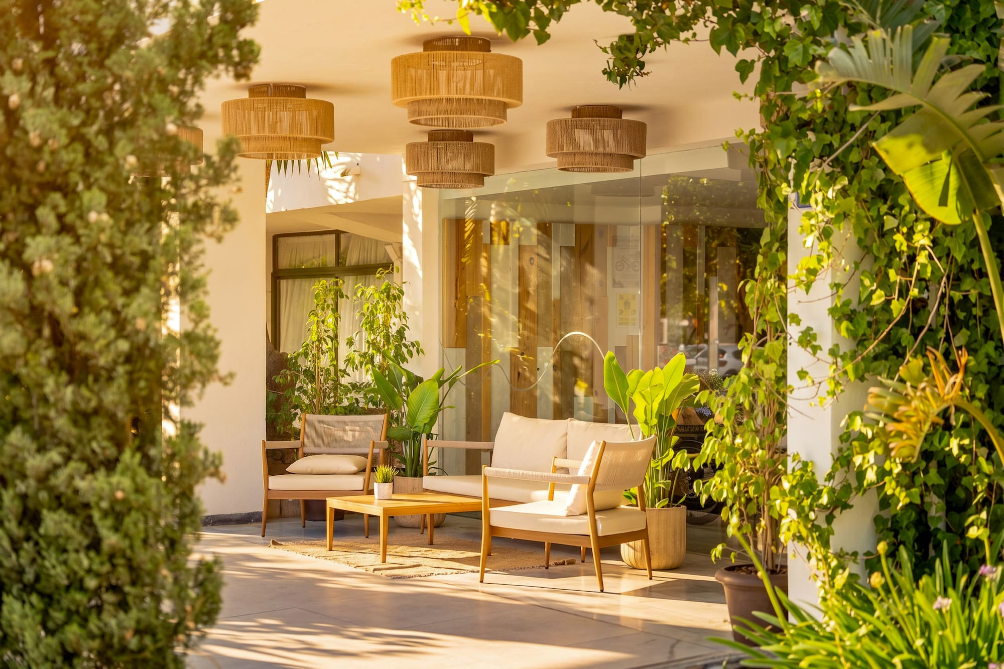 Modern outdoor lounge patio with light furniture, rattan lights, and lush plants.