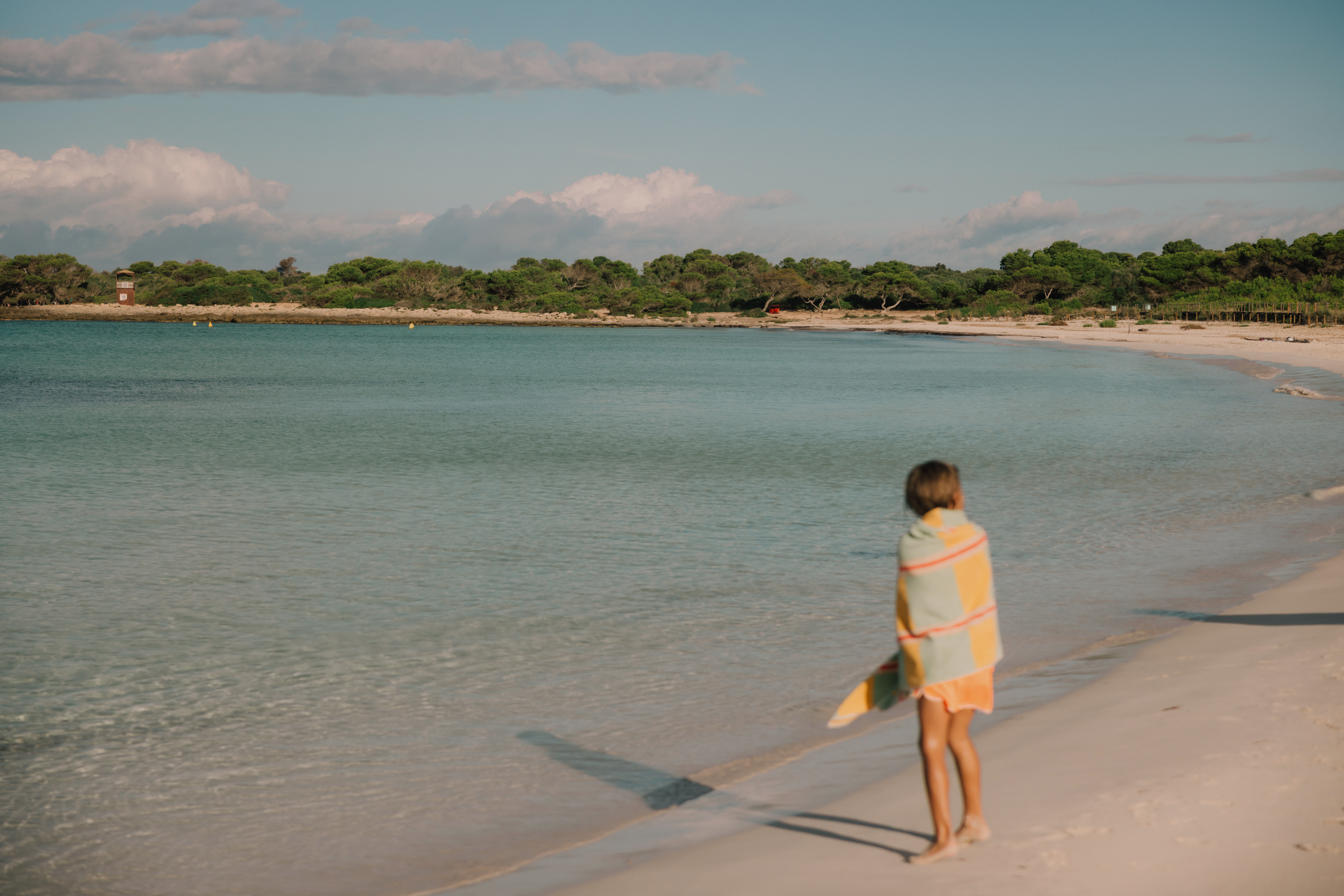 Niño con toalla, playa de arena, agua cristalina y bosque al fondo.