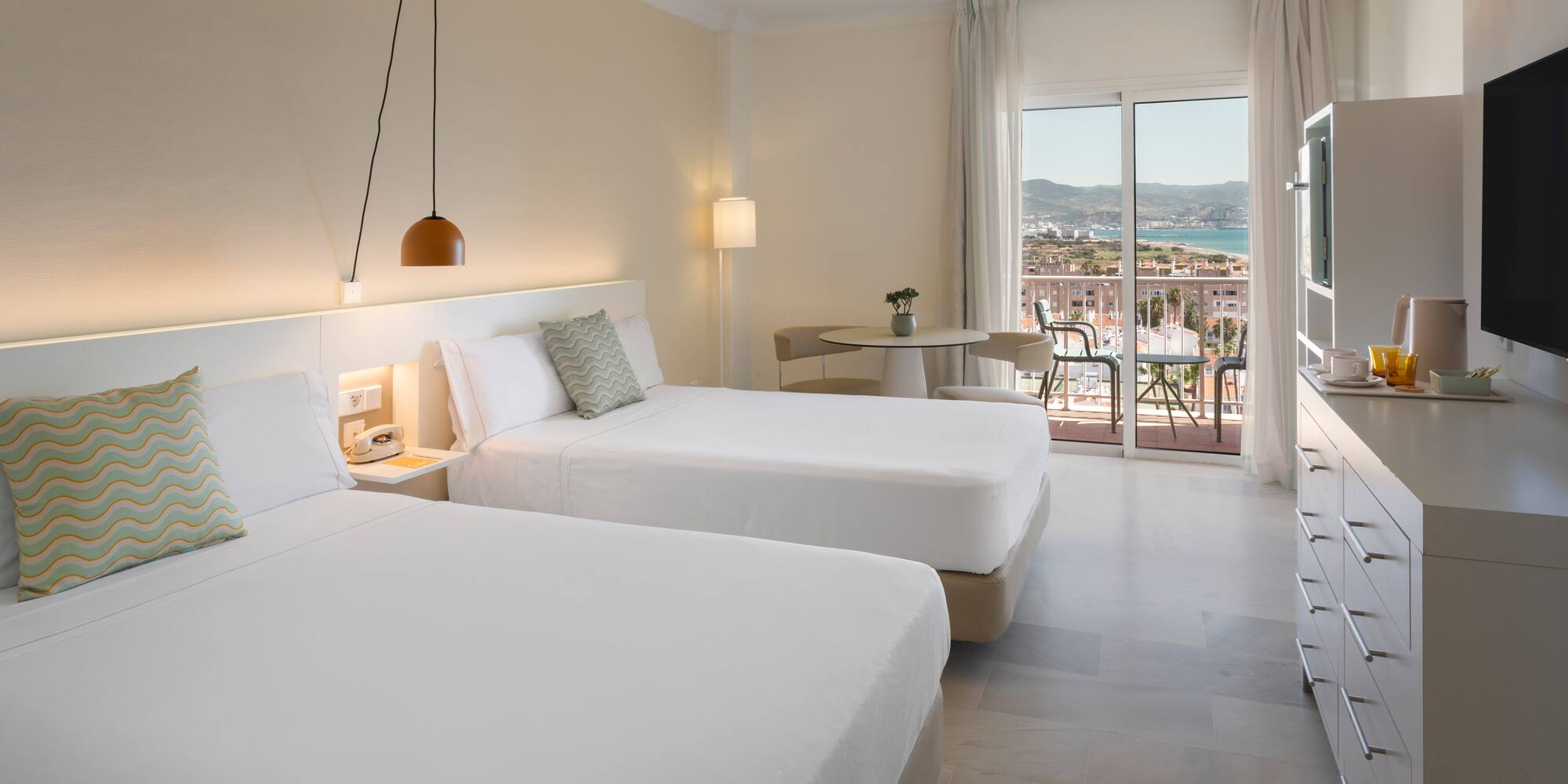 Habitación de hotel con dos camas, balcón y vistas al mar y ciudad.