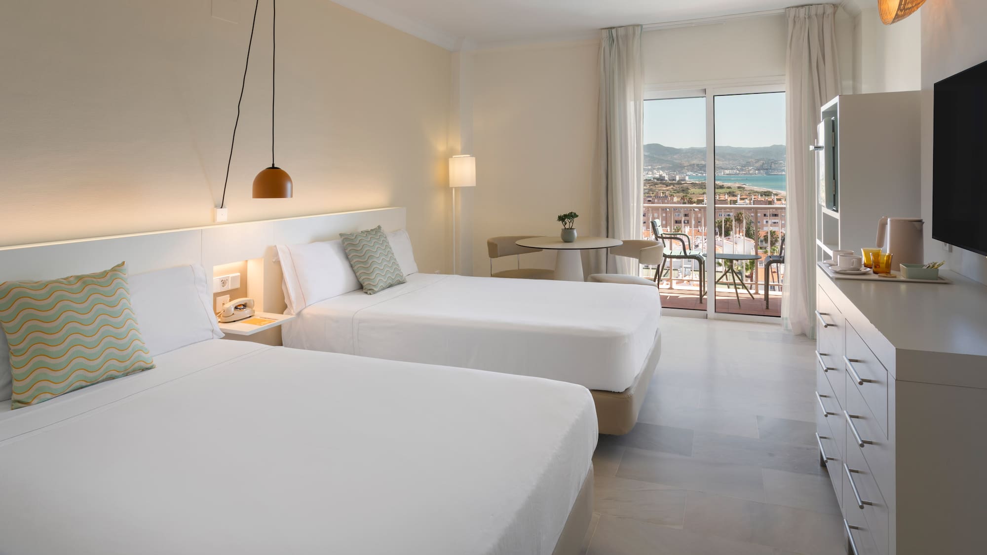 Habitación de hotel con dos camas, balcón y vistas al mar y ciudad.