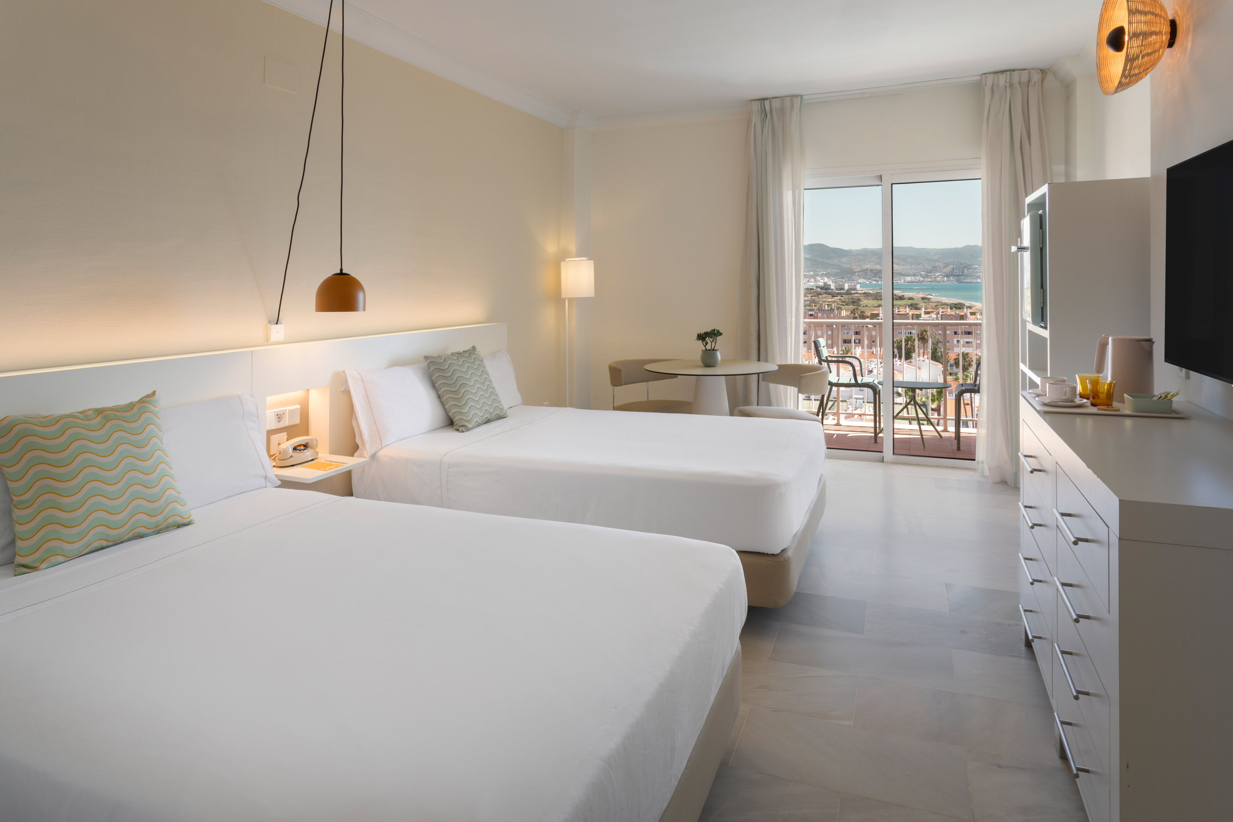 Habitación de hotel con dos camas, balcón y vistas al mar y ciudad.