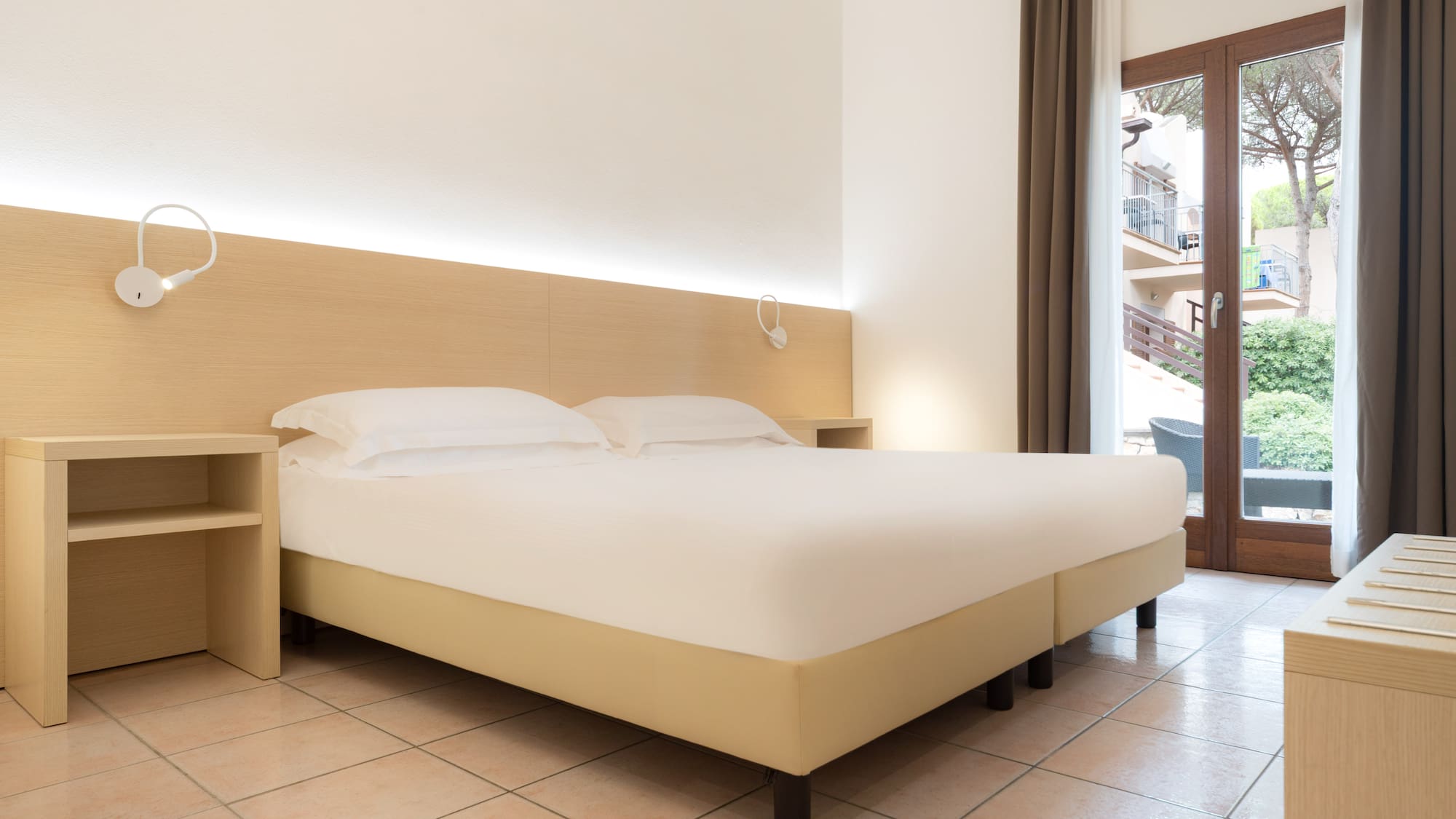 Bright hotel room with double bed, wood headboard, nightstands, balcony view.