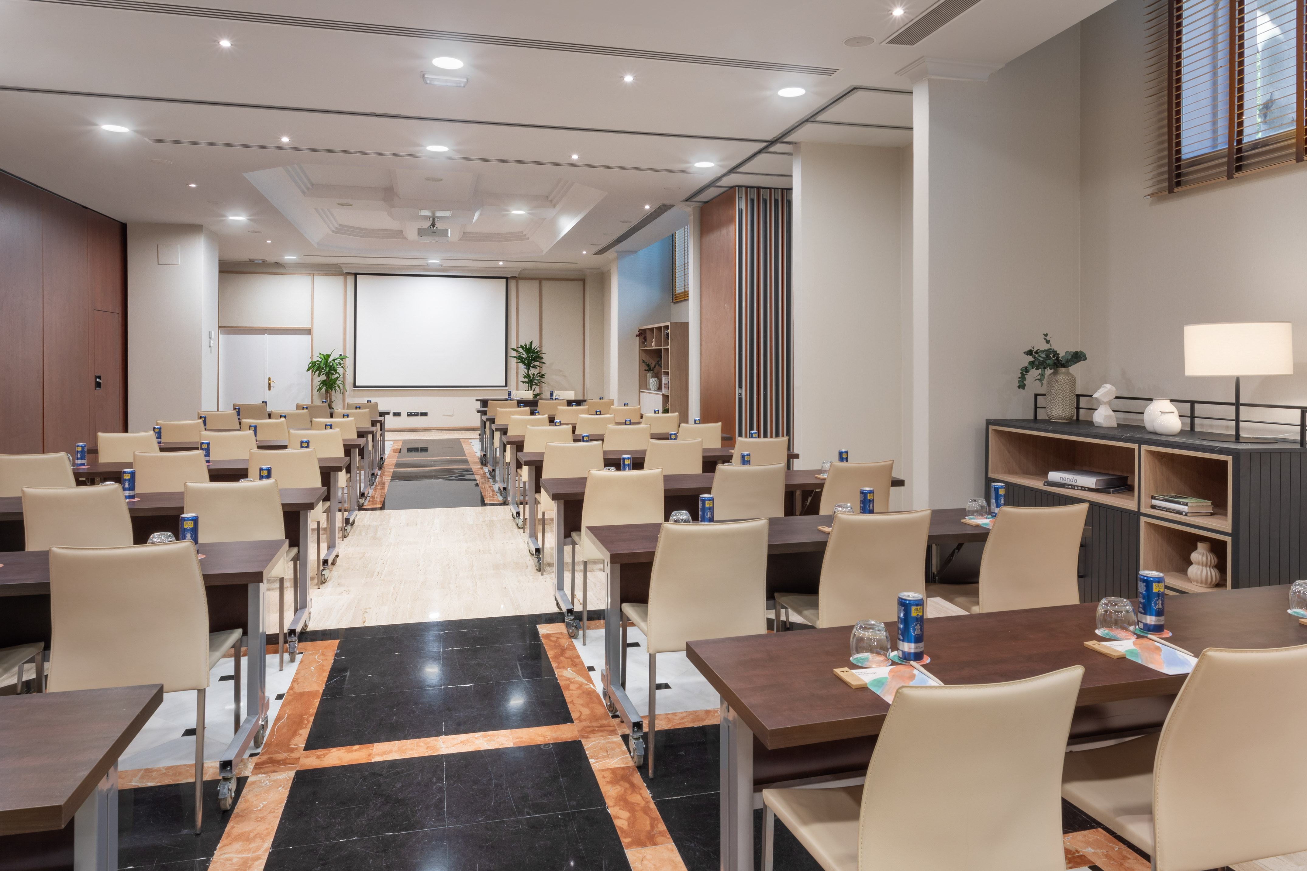 Hotel meeting room with projector screen, rows of tables, and beige chairs.