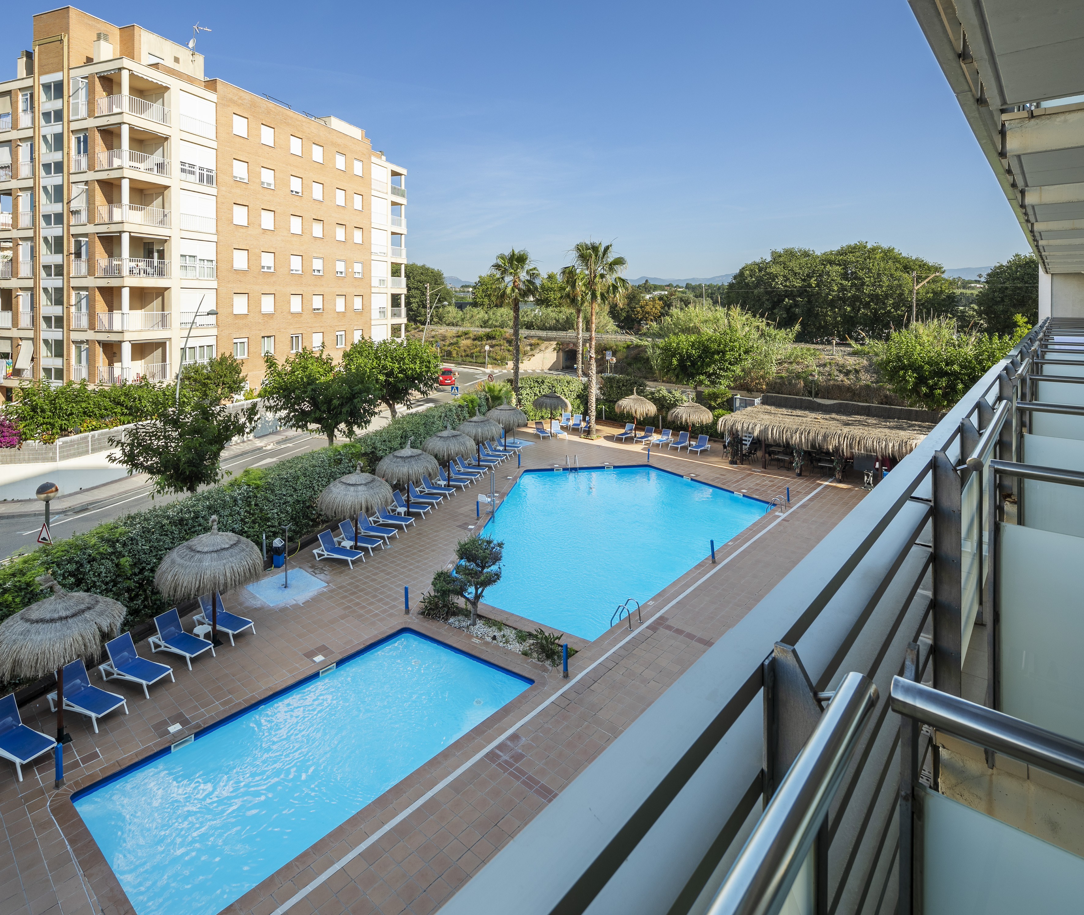 Hotel swimming pools, blue lounge chairs, straw umbrellas, and apartment building.