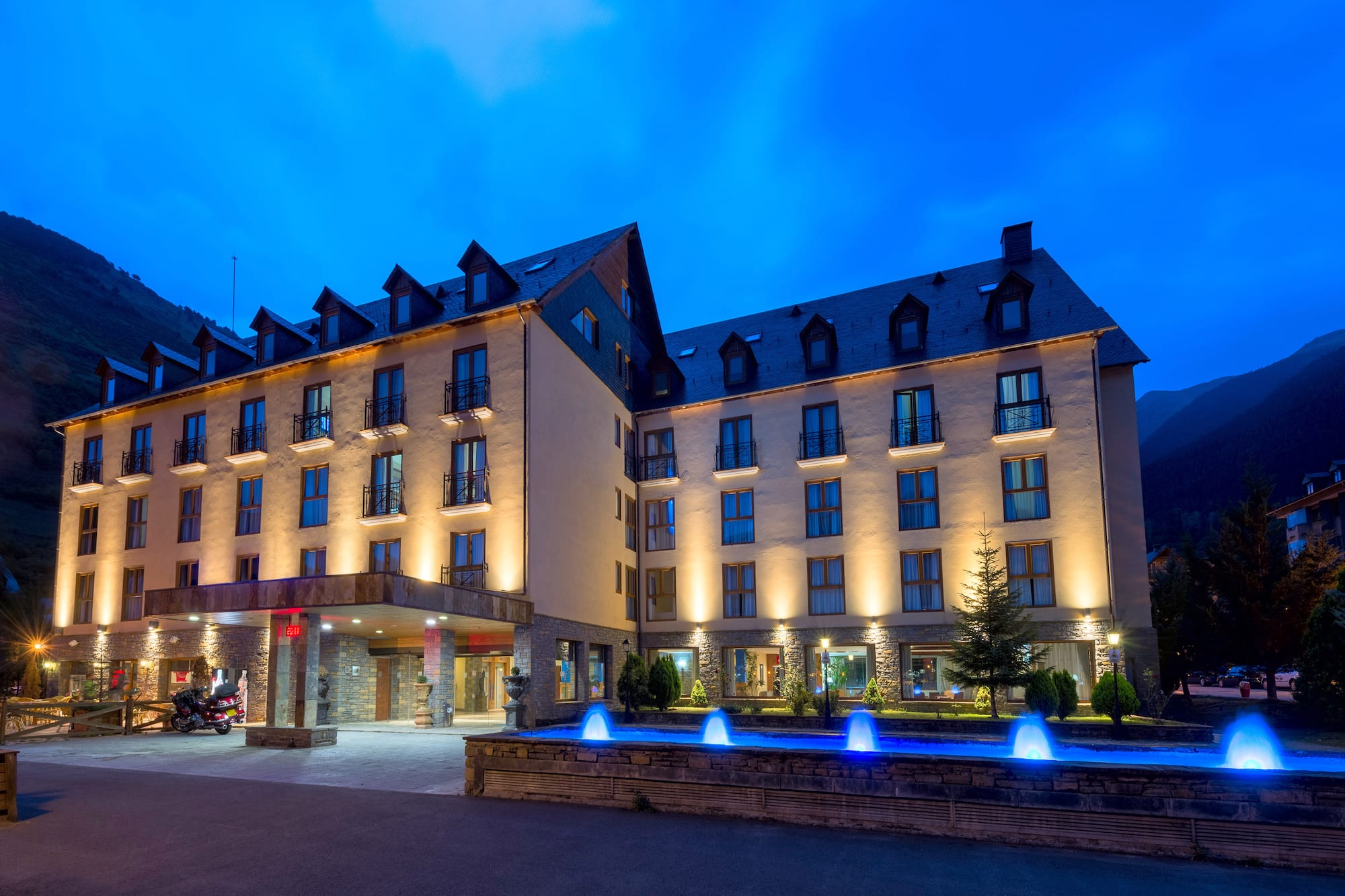 Illuminated mountain hotel exterior at night with blue fountains.