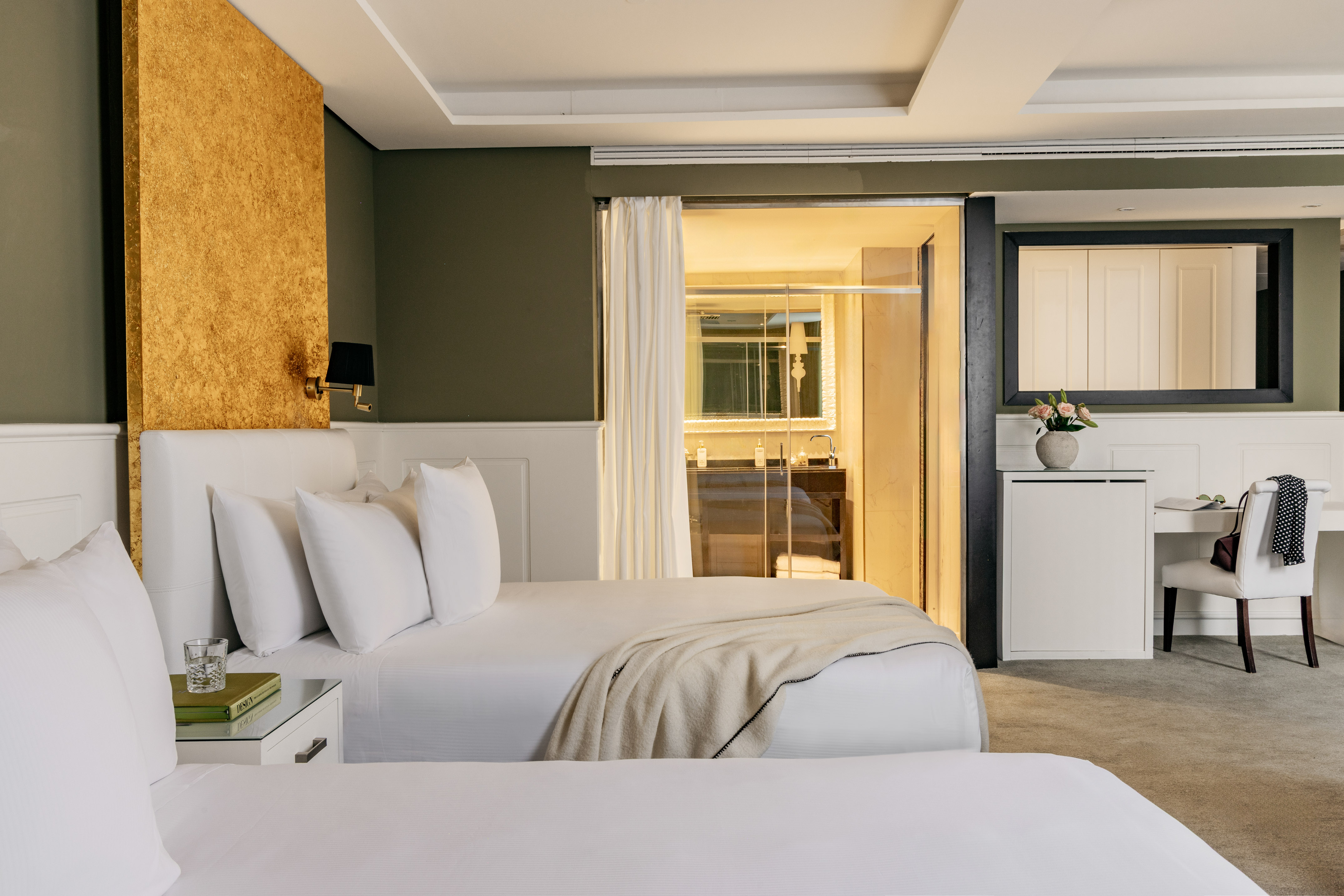 Stylish hotel guest room with twin beds, gold accent wall, and visible bathroom.