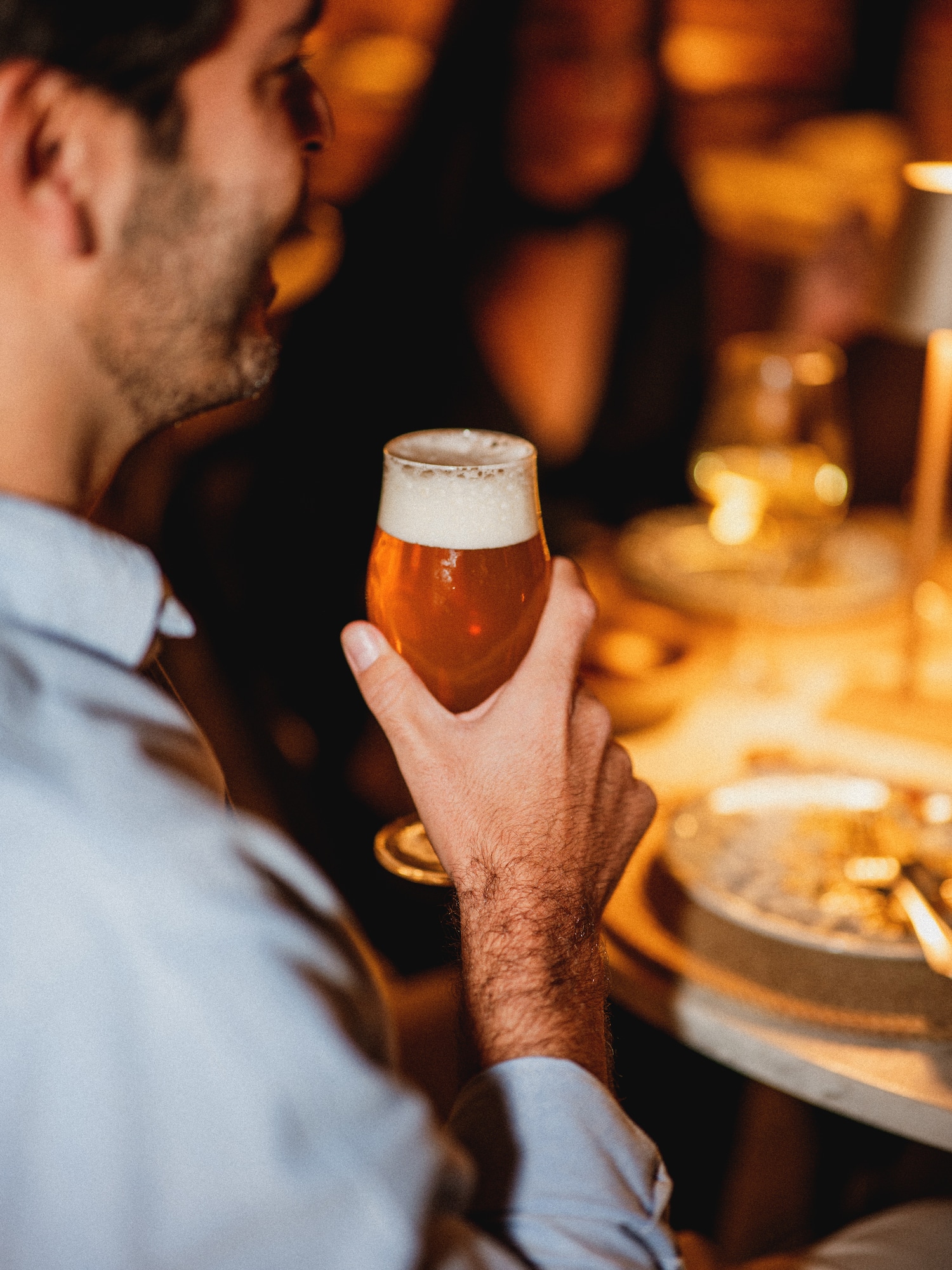 Man holding a glass of amber craft beer with foam at a dim bar or restaurant.