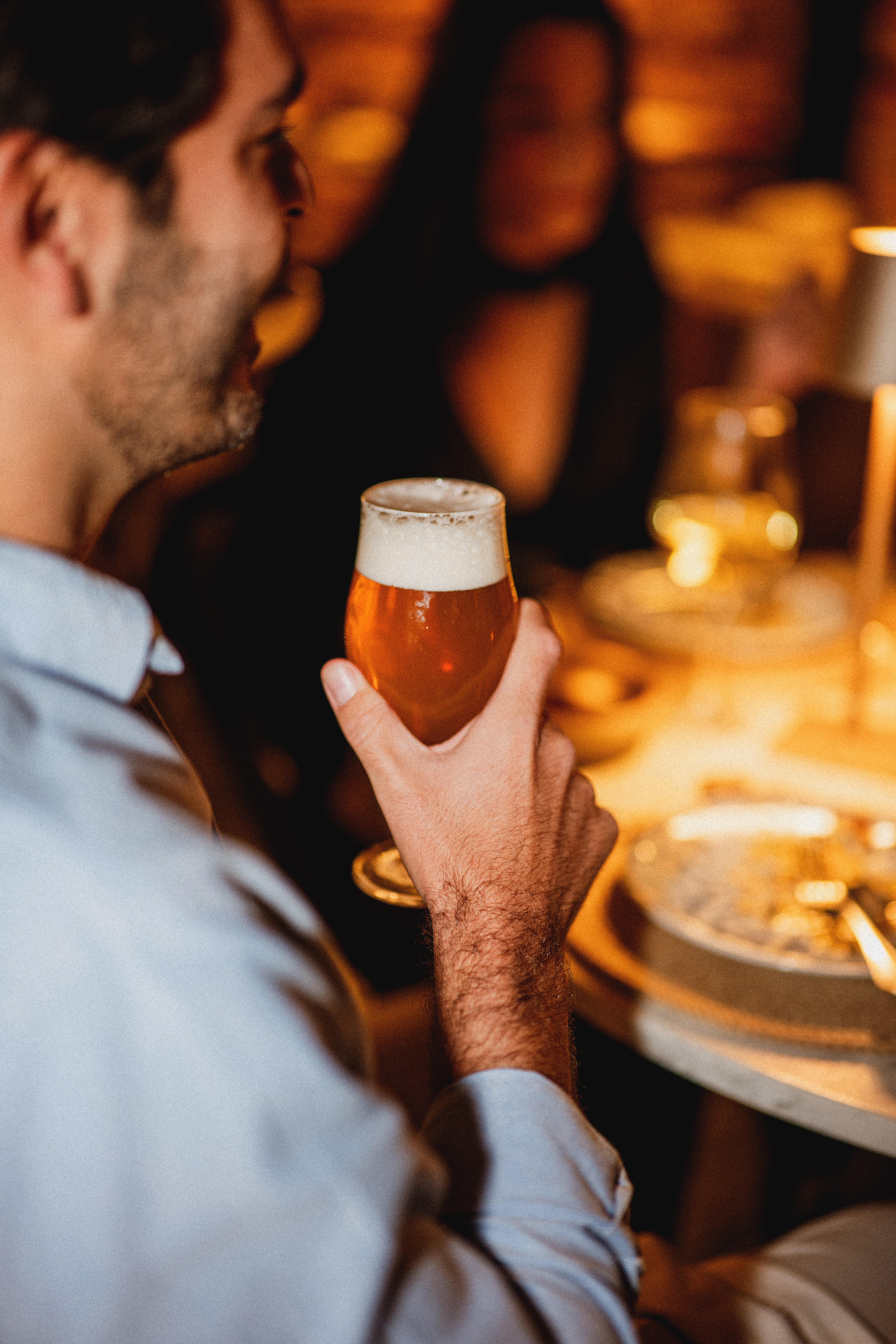 Man holding a glass of amber craft beer with foam at a dim bar or restaurant.