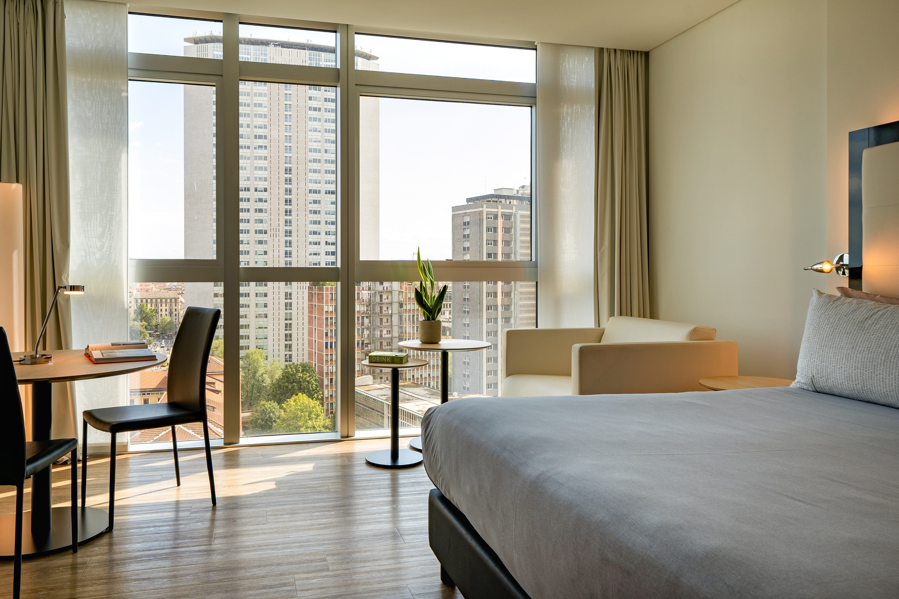 Bright modern hotel room with city skyline view, bed, desk, and armchair.