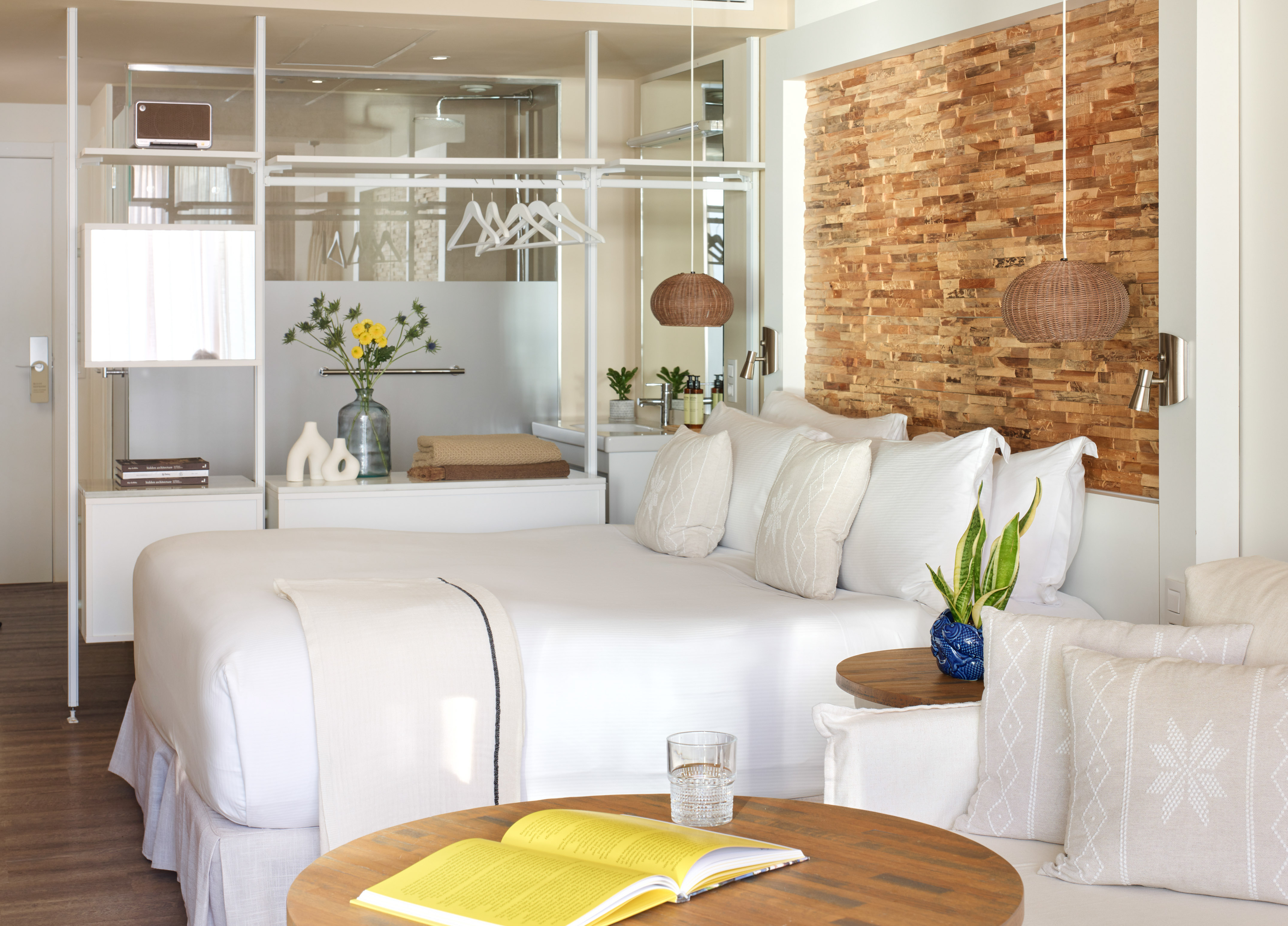 Modern hotel suite with white bed, stone accent wall, and open glass partition.