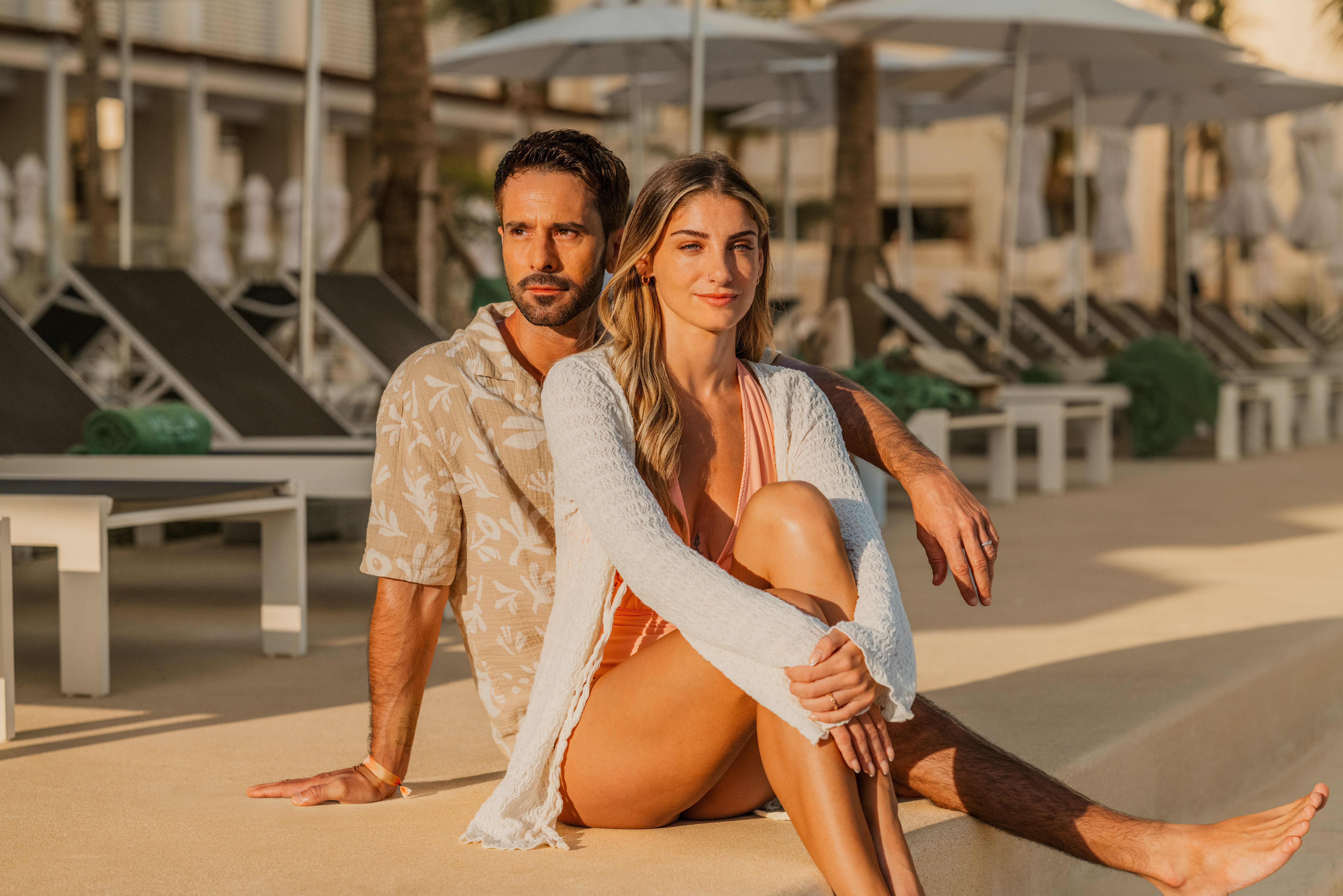 Couple relaxing at sunny resort poolside, lounge chairs in background.