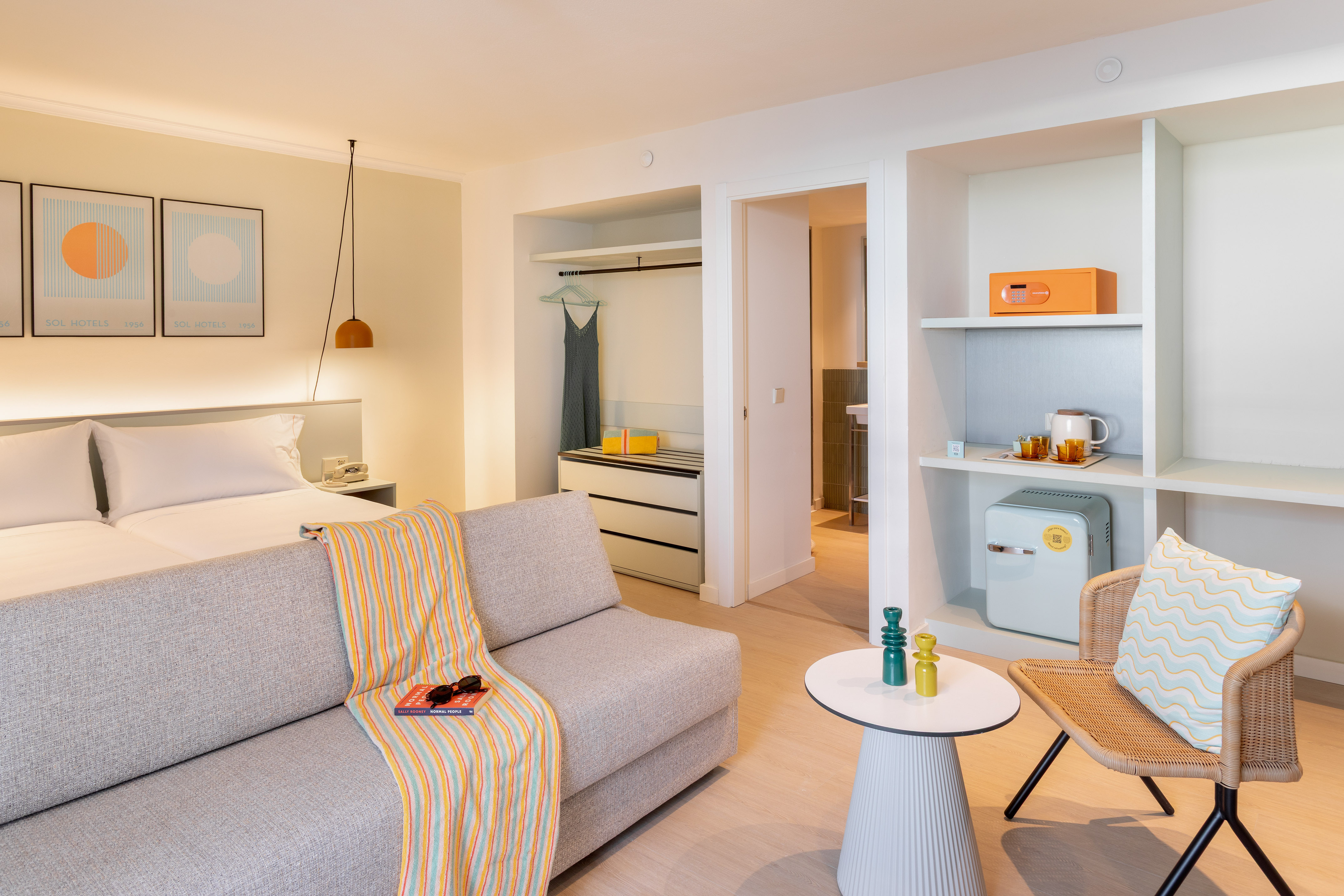 Modern hotel room with twin beds, grey sofa, mini-fridge, and a wicker chair.