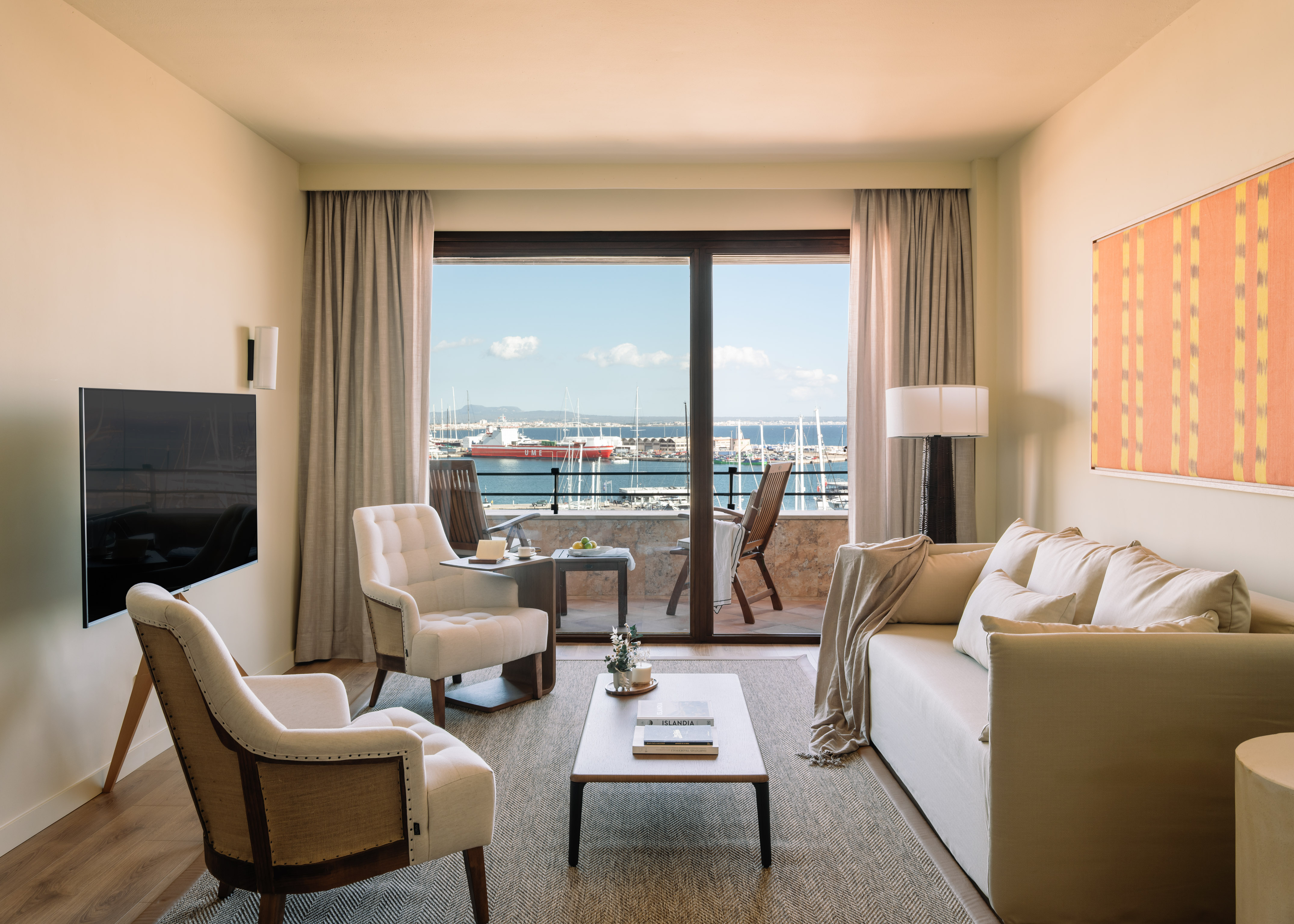 Hotel living room overlooking marina from balcony, sofa, armchairs, and TV.