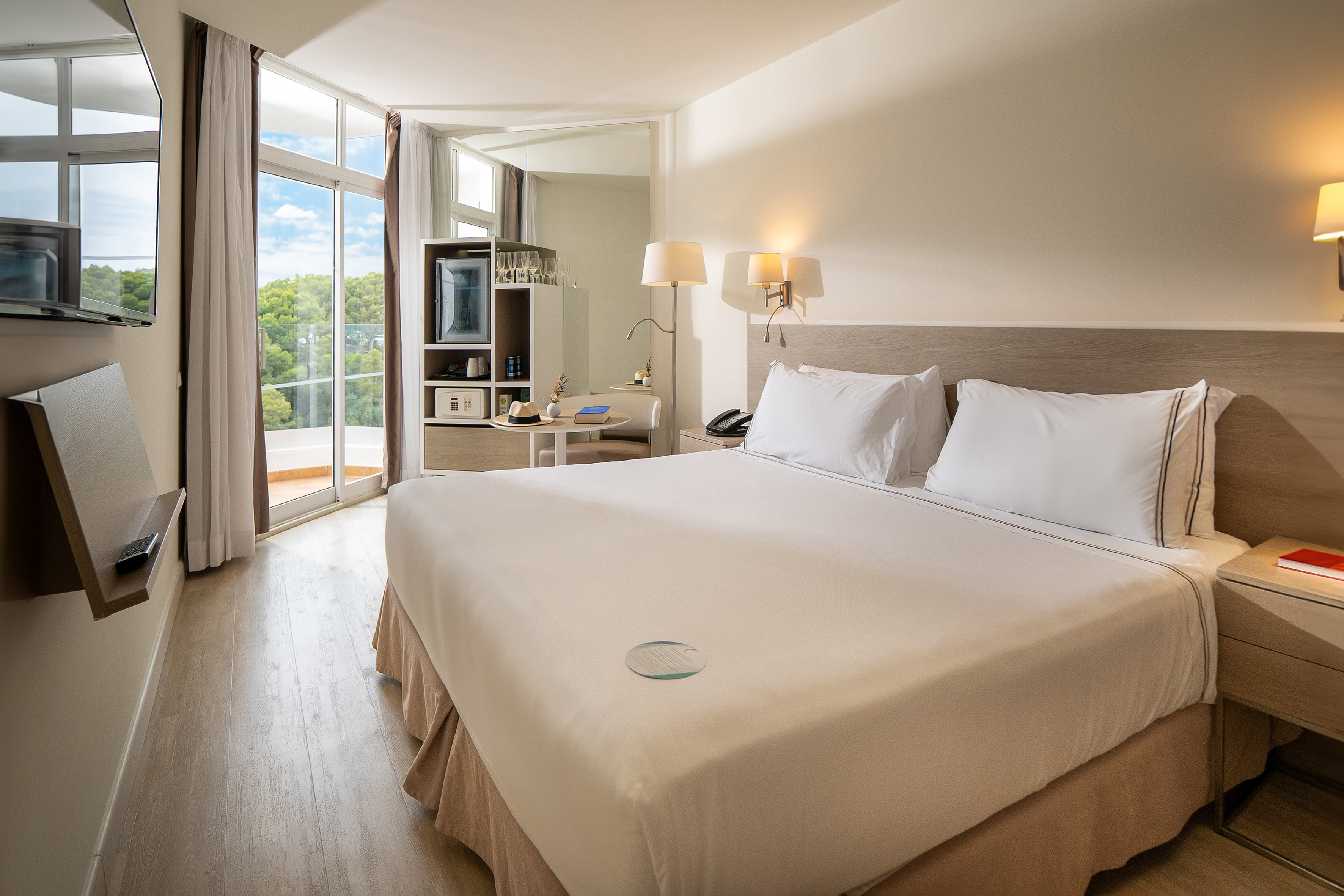Bright hotel room with large bed, balcony, and lush tree view.