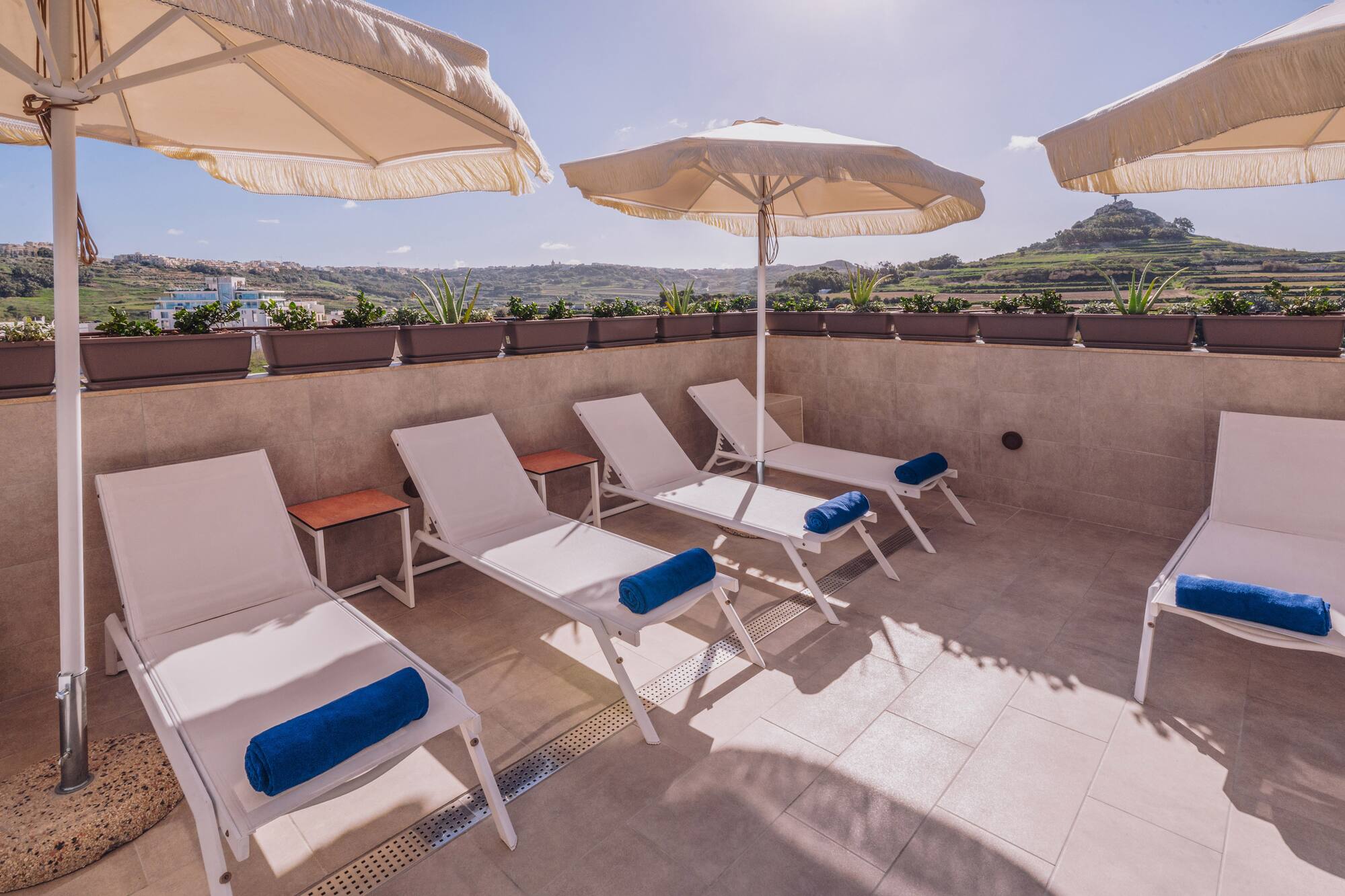 Sunny rooftop terrace with sun loungers, blue towels, and scenic views.