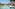Tropical resort pool with modern buildings, palm trees, and sun loungers.