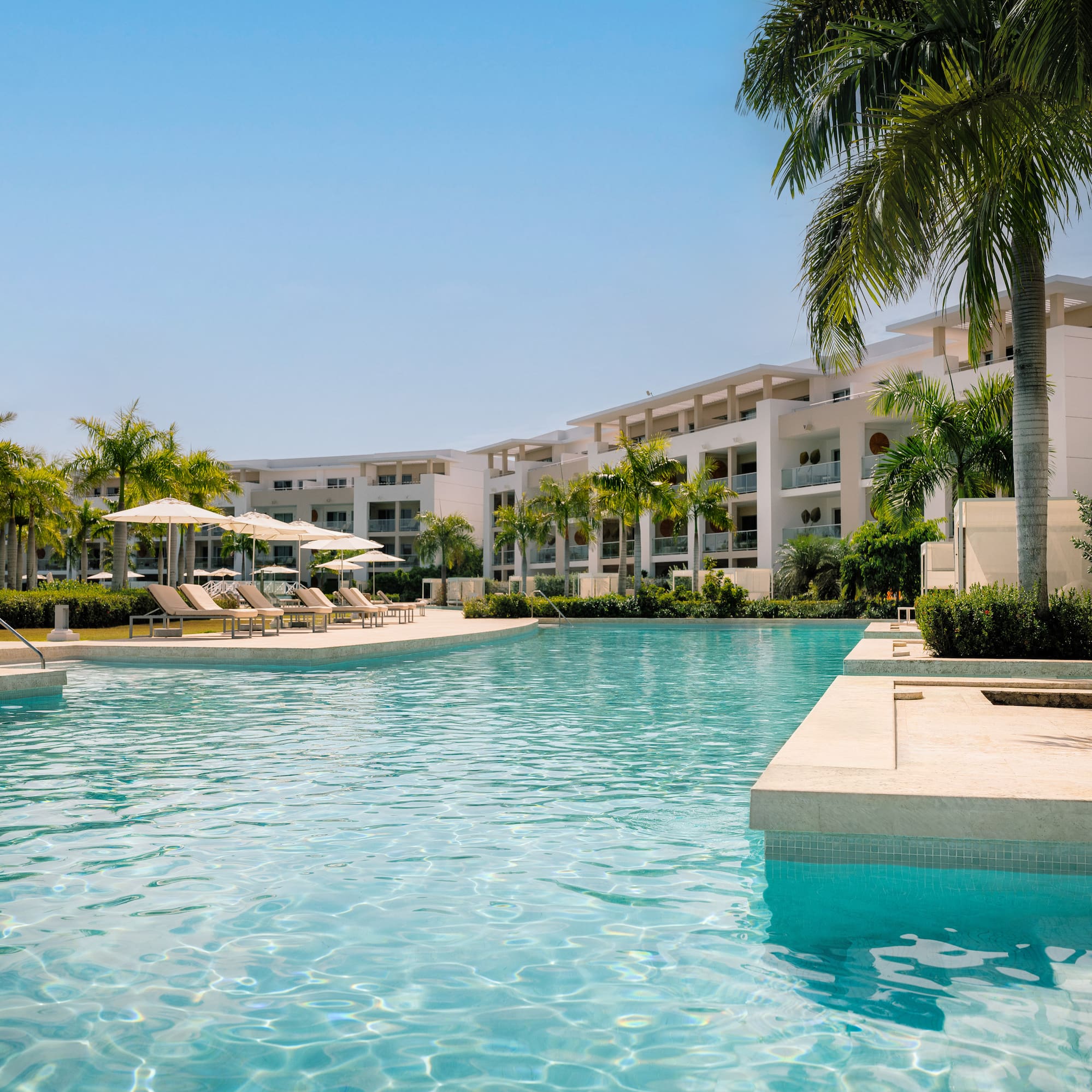 Tropical resort pool with modern buildings, palm trees, and sun loungers.
