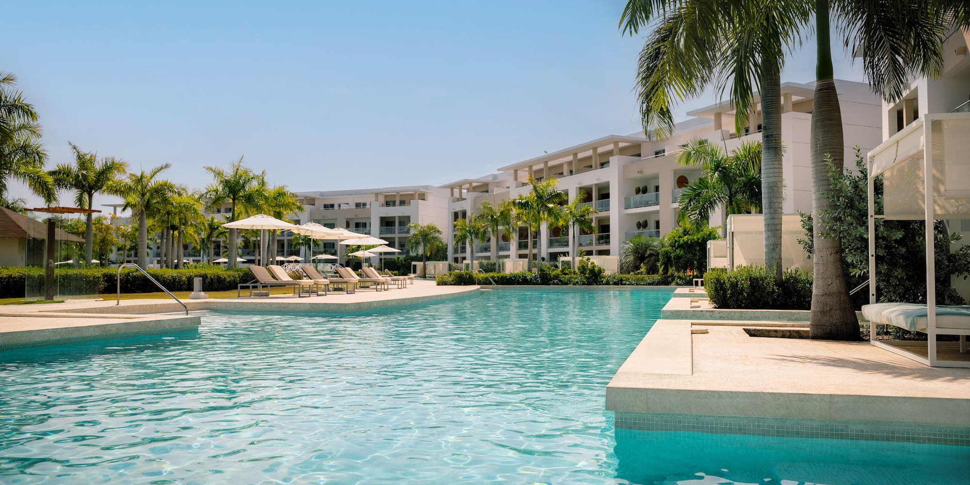 Tropical resort pool with modern buildings, palm trees, and sun loungers.