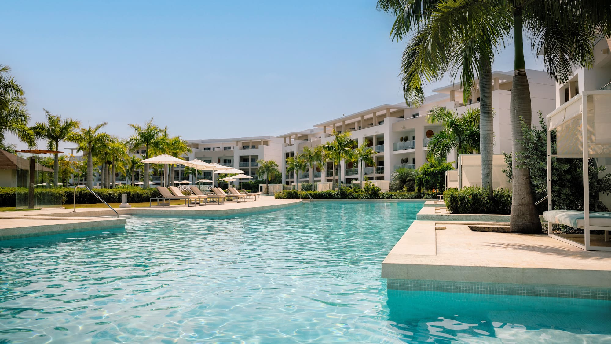 Tropical resort pool with modern buildings, palm trees, and sun loungers.