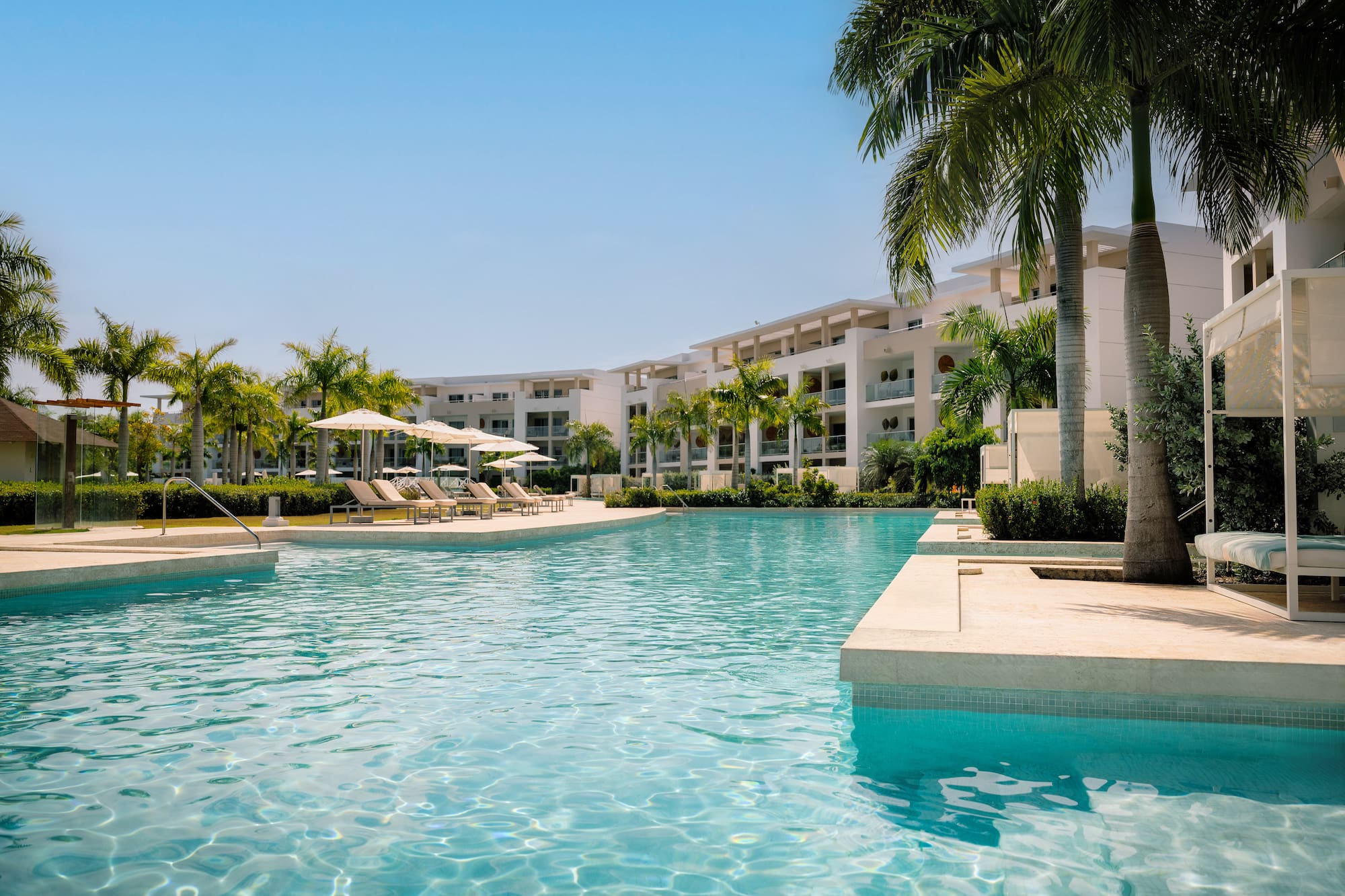 Tropical resort pool with modern buildings, palm trees, and sun loungers.
