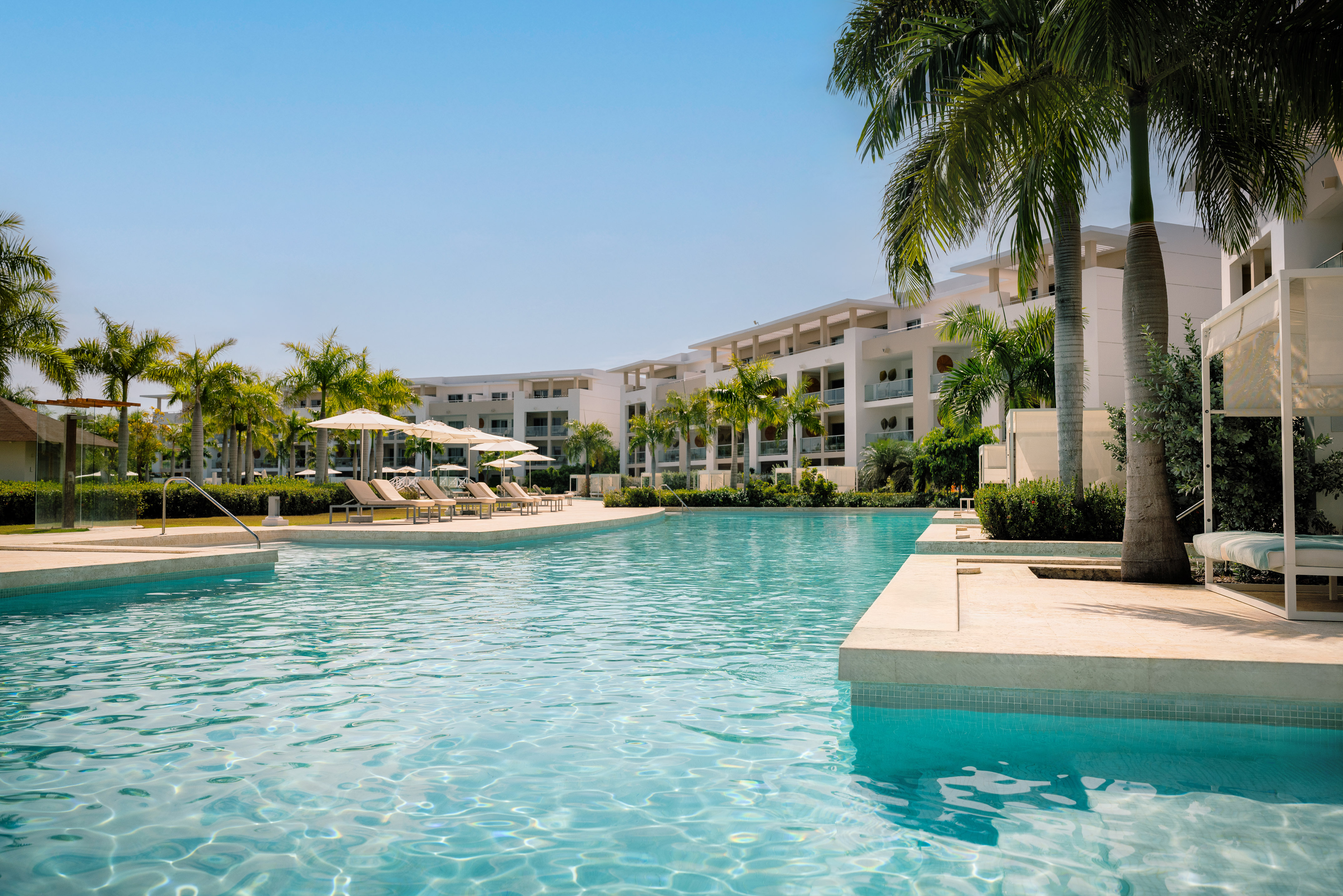 Tropical resort pool with modern buildings, palm trees, and sun loungers.